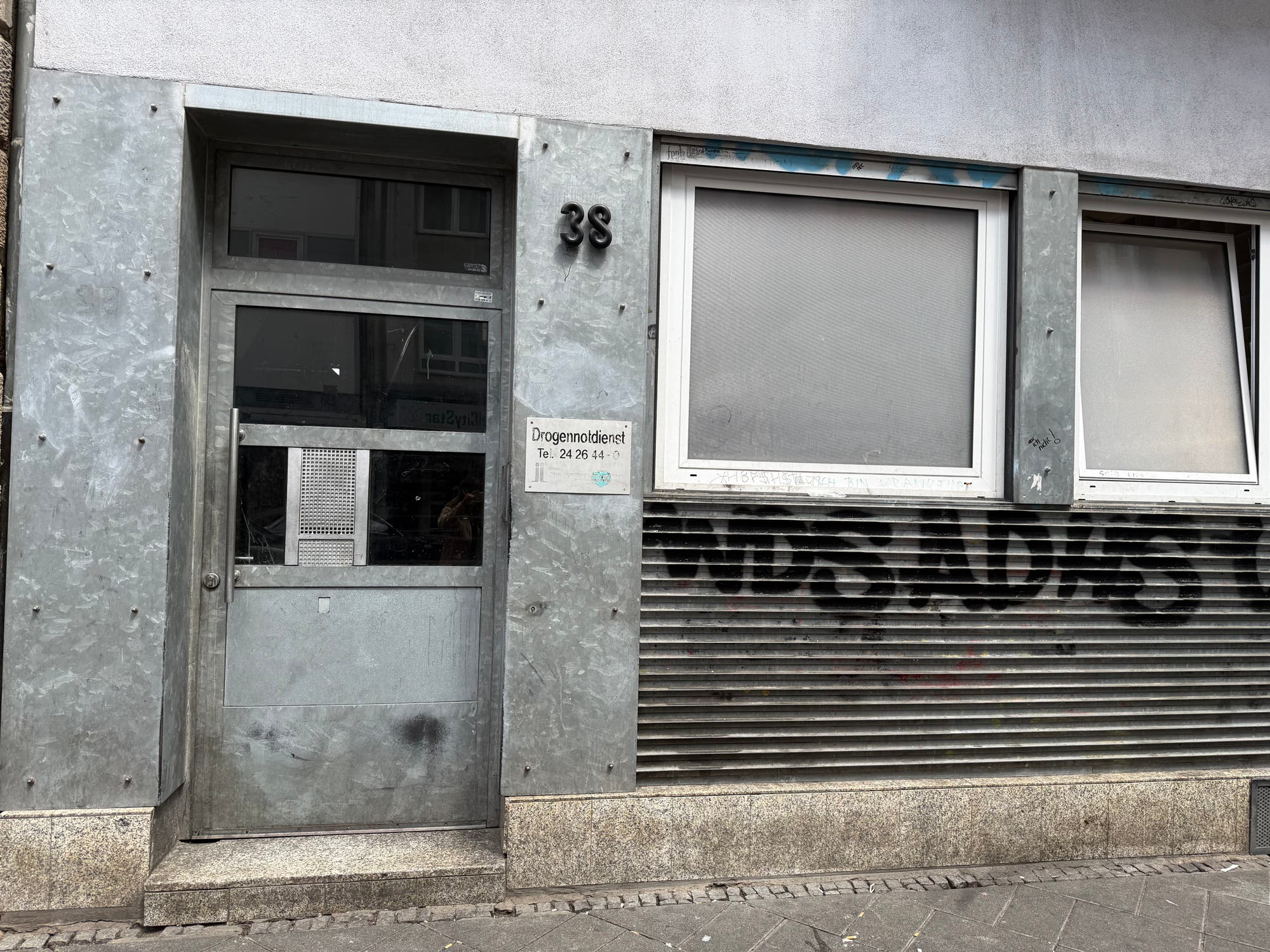 Ein graues Gebäude mit der Hausnummer 38, es wirkt nicht sehr einladend. Links befindet sich eine Metalltür mit einem Klingelschild. Rechts daneben ist ein Fenster mit geschlossenen Jalousien. An der Wand hängt ein Schild mit der Aufschrift "Drogenmoldienst, Tel: 24 26 44-1". Unter dem Fenster ist eine Metallverkleidung mit Graffiti. Die Straße davor ist gepflastert.