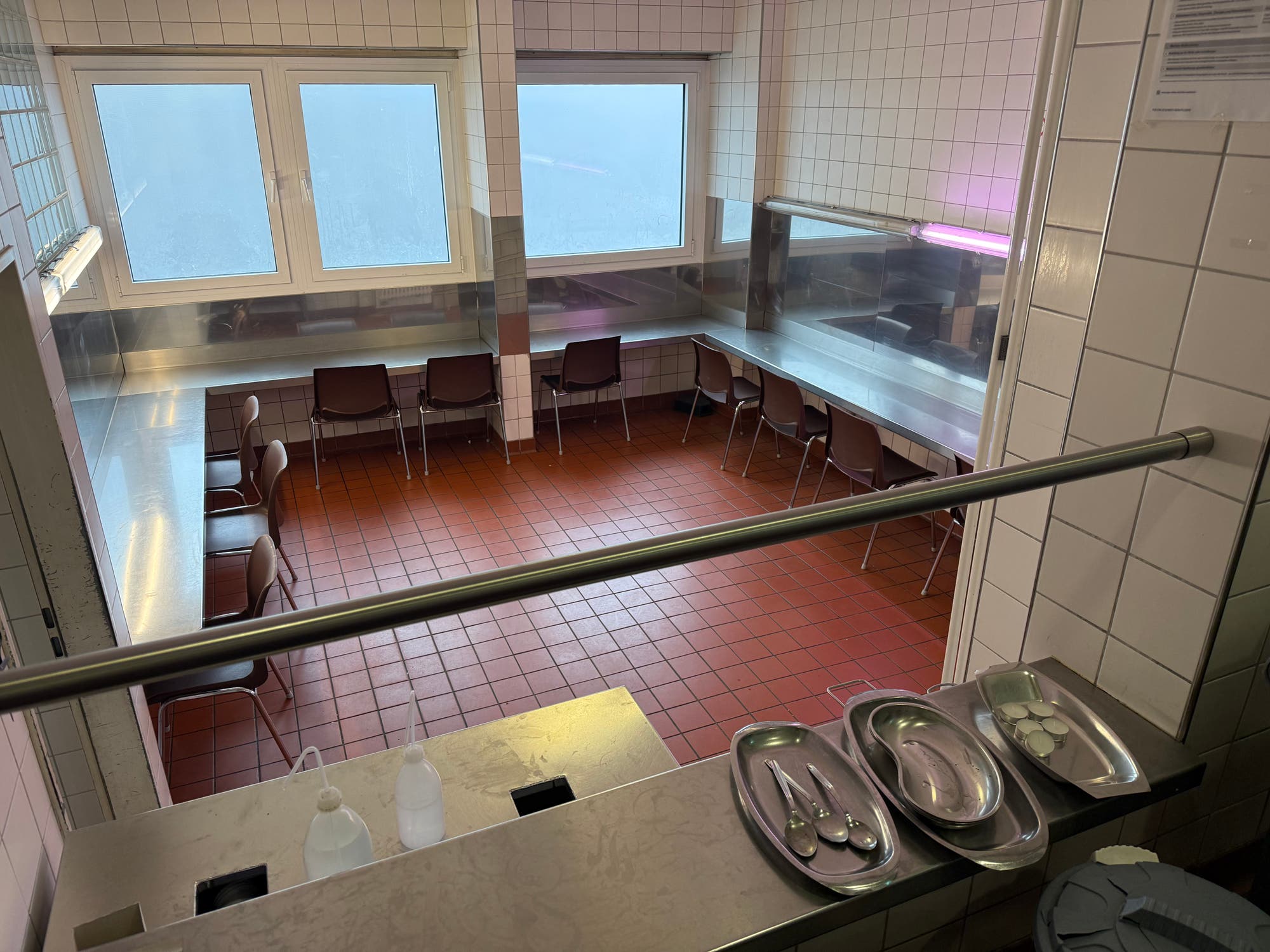 Ein Raum mit gefliesten Wänden und Boden, ausgestattet mit einer U-förmigen Edelstahl-Arbeitsfläche und mehreren Stühlen. Auf der Arbeitsfläche stehen Tabletts mit Löffeln und Tabletten. Im Vordergrund sind Desinfektionsmittelspender zu sehen. Der Raum hat große Fenster, die diffuses Licht hereinlassen.