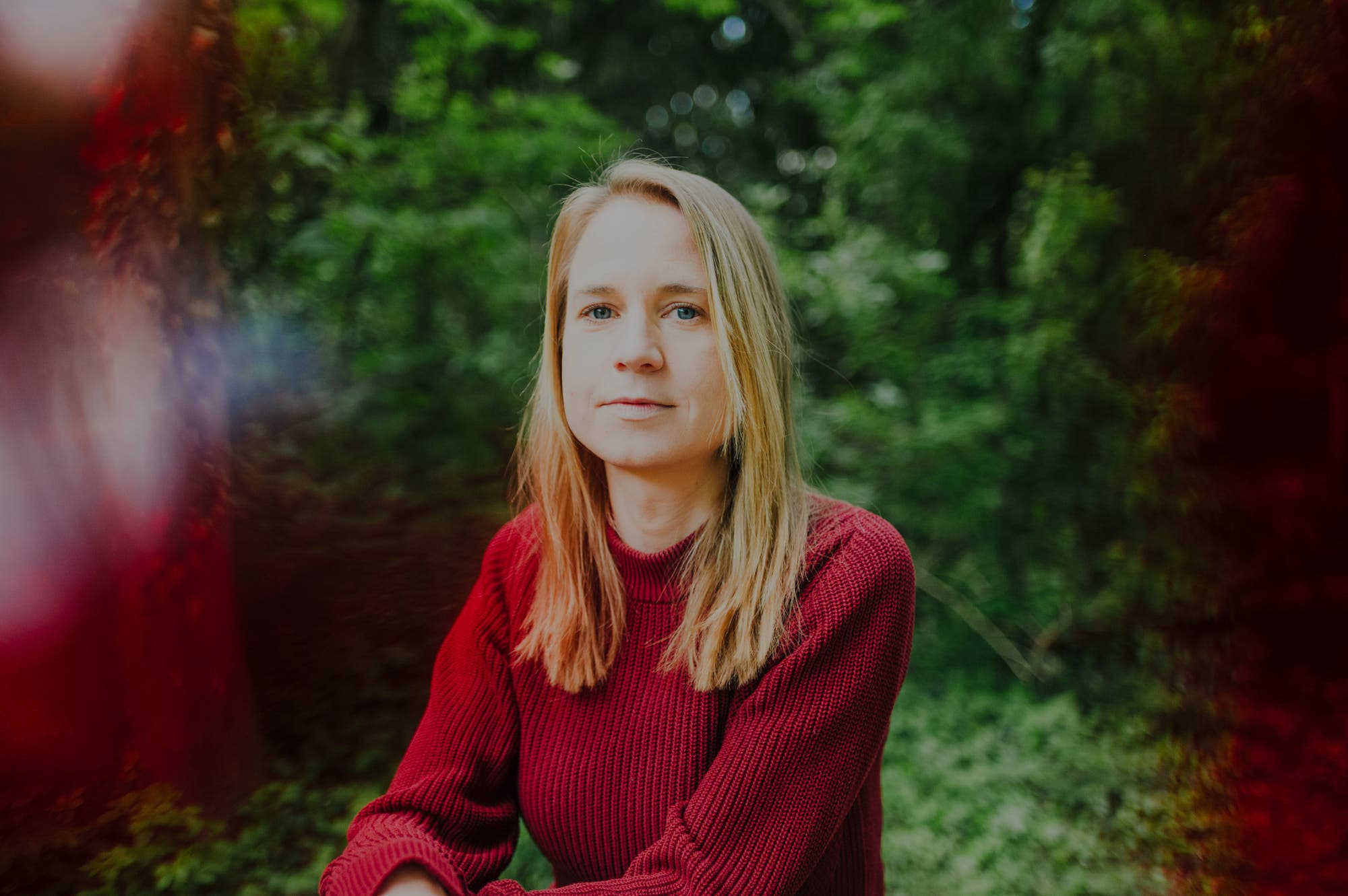 Eine Frau in einem roten Pullover und mit langen hellbrauen Haaren sitzt in einem grünen, bewaldeten Bereich. Die Umgebung ist unscharf, mit einem leichten roten und blauen Schimmer an den Rändern des Bildes. Die Frau blickt ruhig in die Kamera.