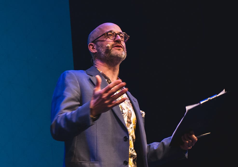 Eine Person in einem blauen Anzug hält eine Präsentation auf einer Bühne. Sie trägt ein Headset-Mikrofon und hält in einer Hand ein Klemmbrett. Der Hintergrund ist dunkel, mit einem blauen Bereich auf der linken Seite. Die Person gestikuliert mit der freien Hand, als ob sie einen Punkt betont.