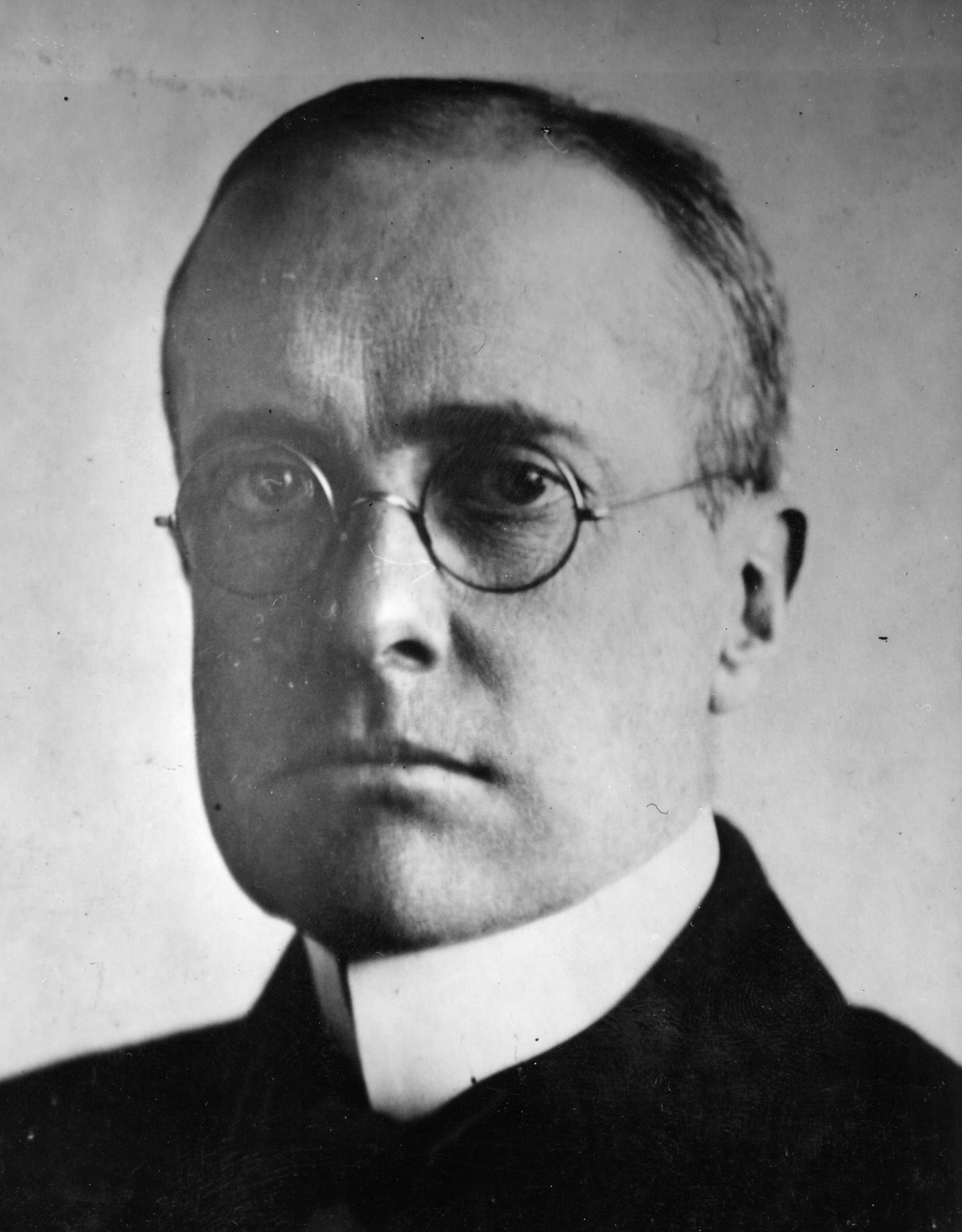 Ein altes Schwarz-Weiß-Foto eines Mannes in Hemd und Anzug, der ernst in die Kamera blickt. Er trägt eine Nickelbrille und keinen Bart. Die Haare sind ordentlich eng an den Kopf frisiert. Der Hintergrund ist neutral und schlicht.