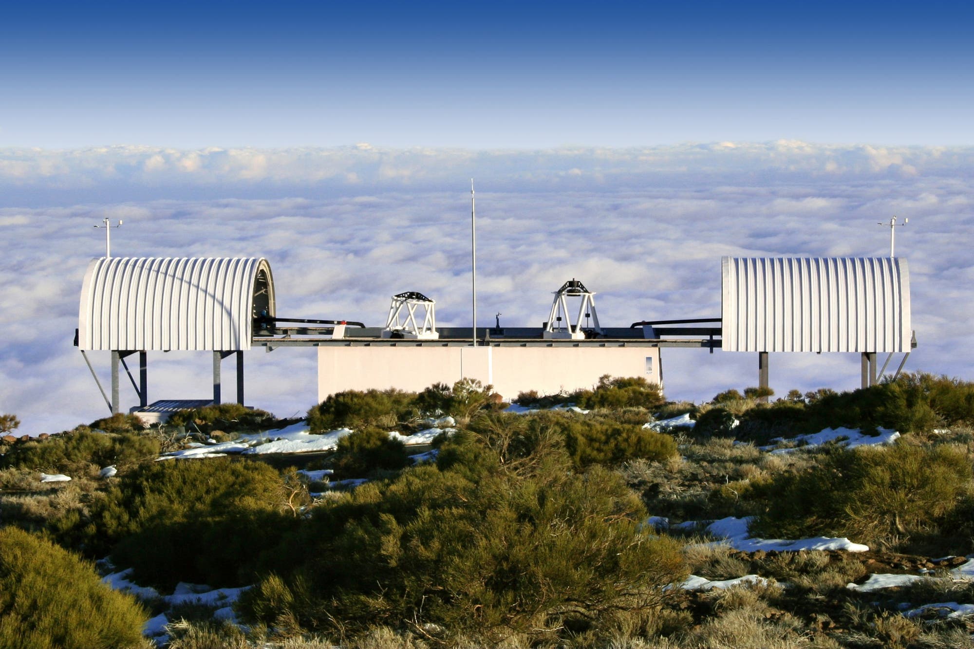 Ein Observatorium mit zwei großen, gewölbten Dächern steht auf einer Anhöhe über einer Wolkendecke. Die Struktur ist von niedriger Vegetation und vereinzelten Schneeflecken umgeben. Der Himmel ist klar und blau. 
