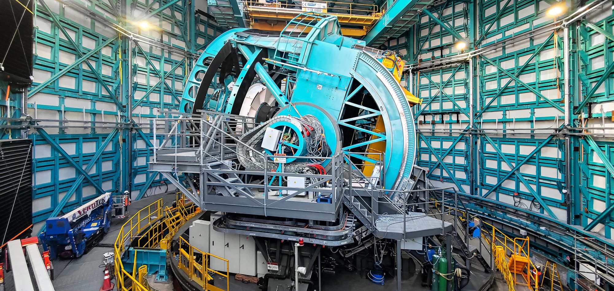 Un telescopio grande y moderno en una cúpula del Observatorio. La estructura está rodeada por una compleja red de plataformas y barandas de metal. El telescopio está pintado en un azul y rodeado de dispositivos y cables técnicos. Las paredes del domo están reforzadas con portadores de metal verde. Ninguna gente es visible.