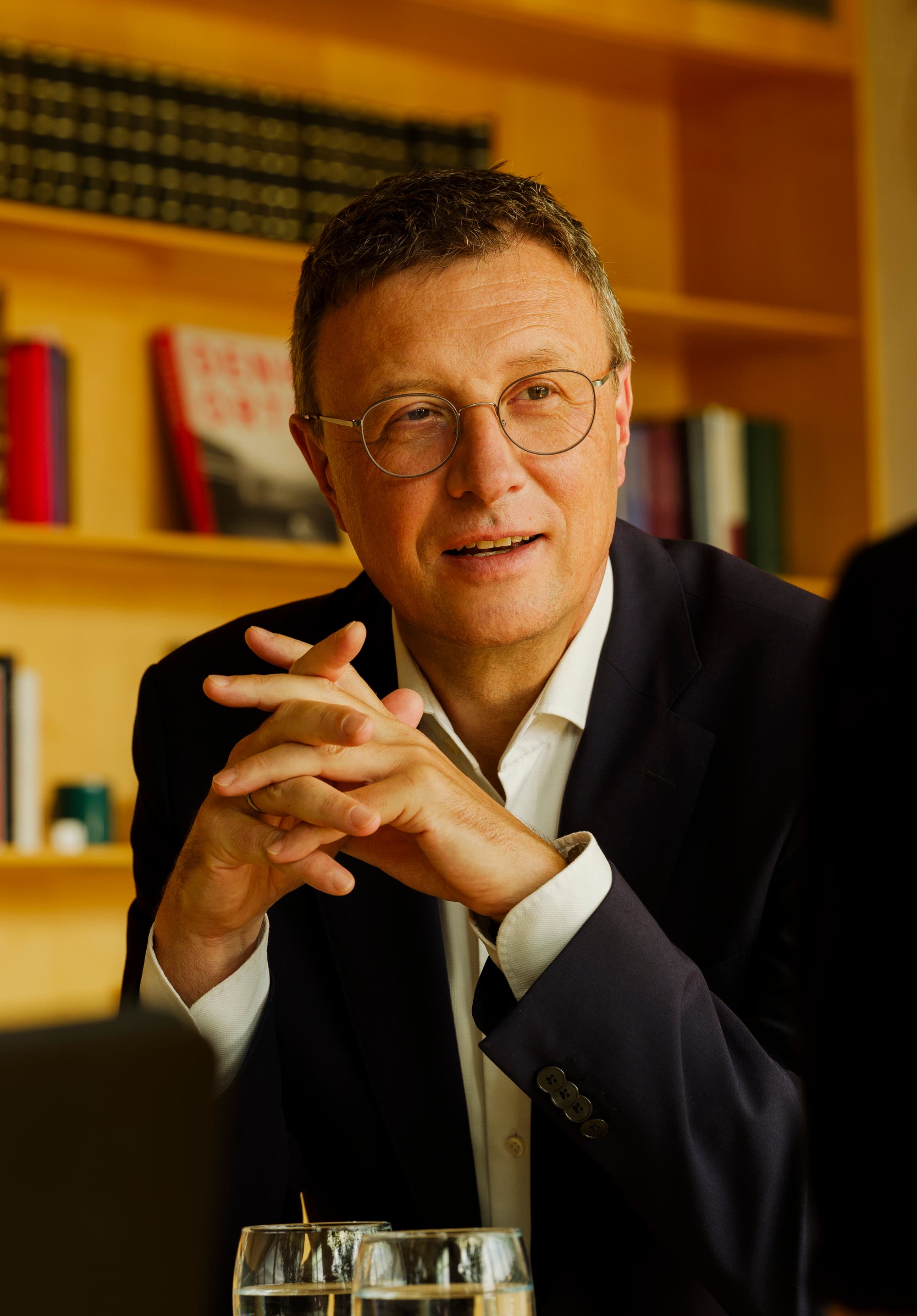 Ein Mann in einem dunklen Anzug steht an einem Tisch mit Bücherregalen im Hintergrund. Er trägt eine Brille und hat die Hände locker vor sich gefaltet. Er spricht und schaut dabei aufmerksam. Auf dem Tisch stehen zwei Gläser mit Wasser. Die Szene vermittelt eine professionelle und konzentrierte Atmosphäre.