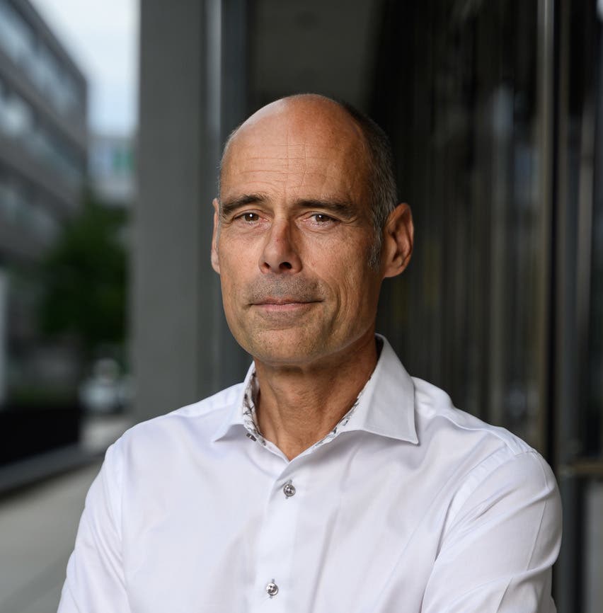 Eine Person in einem weißen Hemd steht draußen vor einem modernen Gebäude. Die Person schaut direkt in die Kamera und hat einen neutralen Gesichtsausdruck. Im Hintergrund sind unscharfe Bürogebäude und ein Gehweg zu sehen.