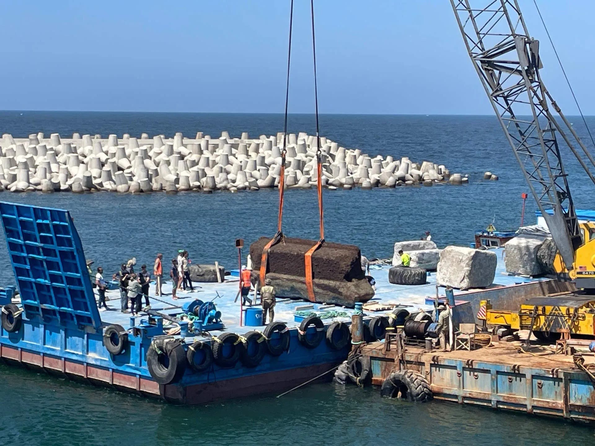 Am Meer steht einen Kran, der einen großen Steinblock auf ein blaues Lastkahn-Deck hebt. Mehrere Arbeiter in Schutzkleidung stehen auf dem Deck und beobachten den Vorgang. Im Hintergrund ist eine Reihe von Wellenbrechern aus Beton zu sehen, die das Meer vom Hafenbereich trennen. Der Himmel ist klar und blau.