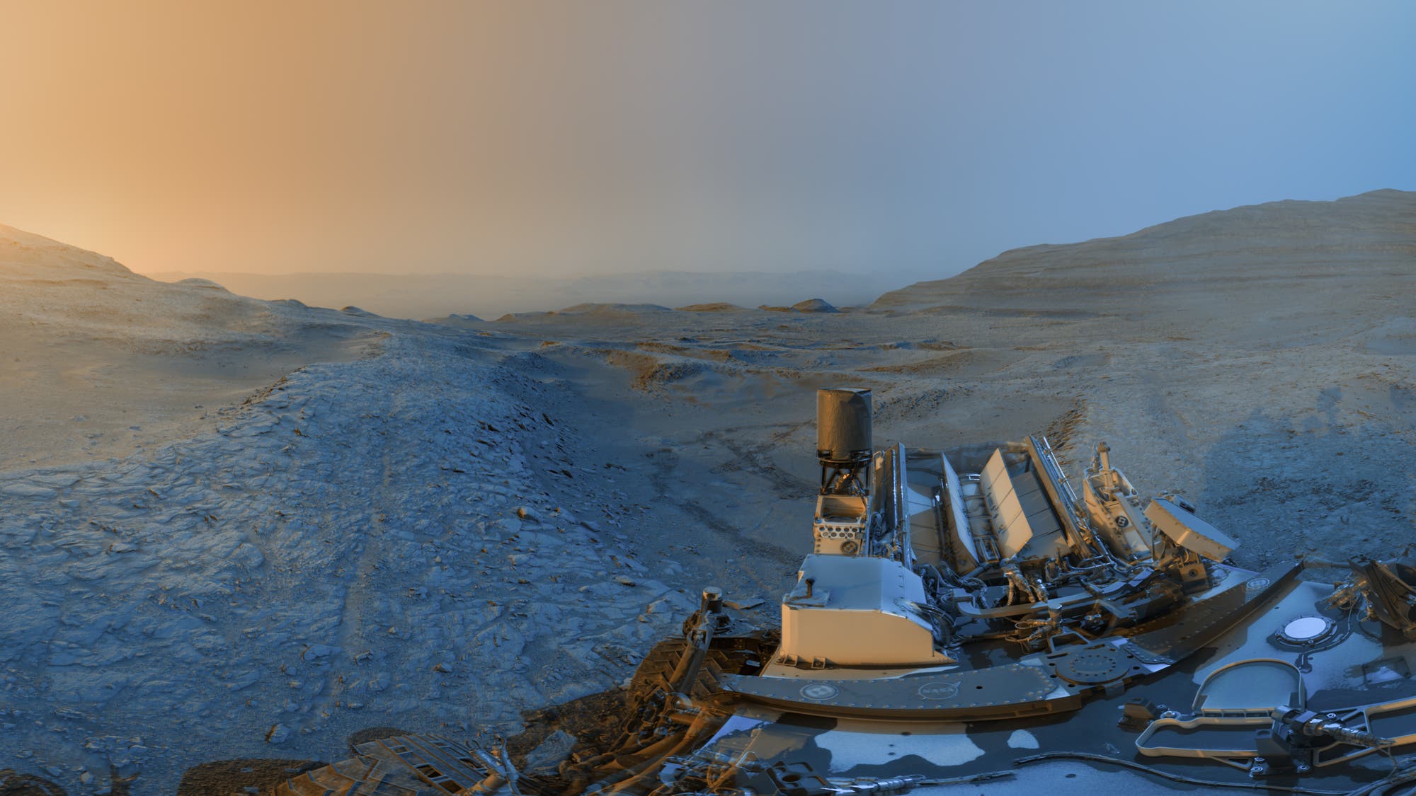 Eine Panoramaaufnahme der Marsoberfl&auml;che, aufgenommen von einem Rover. Im Vordergrund sind Teile des Rovers sichtbar, w&auml;hrend sich im Hintergrund eine weite, felsige Landschaft erstreckt. Der Himmel zeigt einen Farbverlauf von orange zu blau, was auf einen Sonnenaufgang oder Sonnenuntergang hindeutet. Die Szene vermittelt den Eindruck von Ruhe und Weite auf dem Mars.