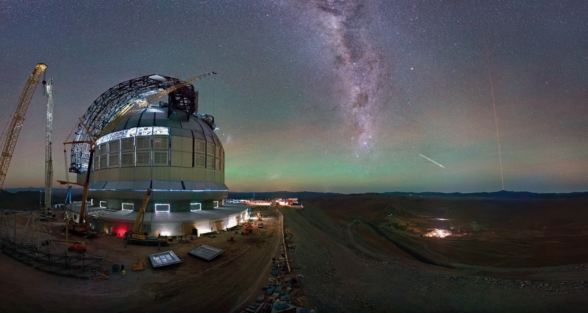 Ein großes Observatorium mit einer Kuppel und Baukränen steht in einer Wüstenlandschaft unter einem klaren Nachthimmel. Die Milchstraße ist deutlich sichtbar, und ein heller Meteor zieht eine Linie am Himmel. Im Hintergrund sind Berge und Lichter einer entfernten Siedlung zu sehen. Die Szene vermittelt eine ruhige, aber beeindruckende Atmosphäre der Astronomie und Technik.
