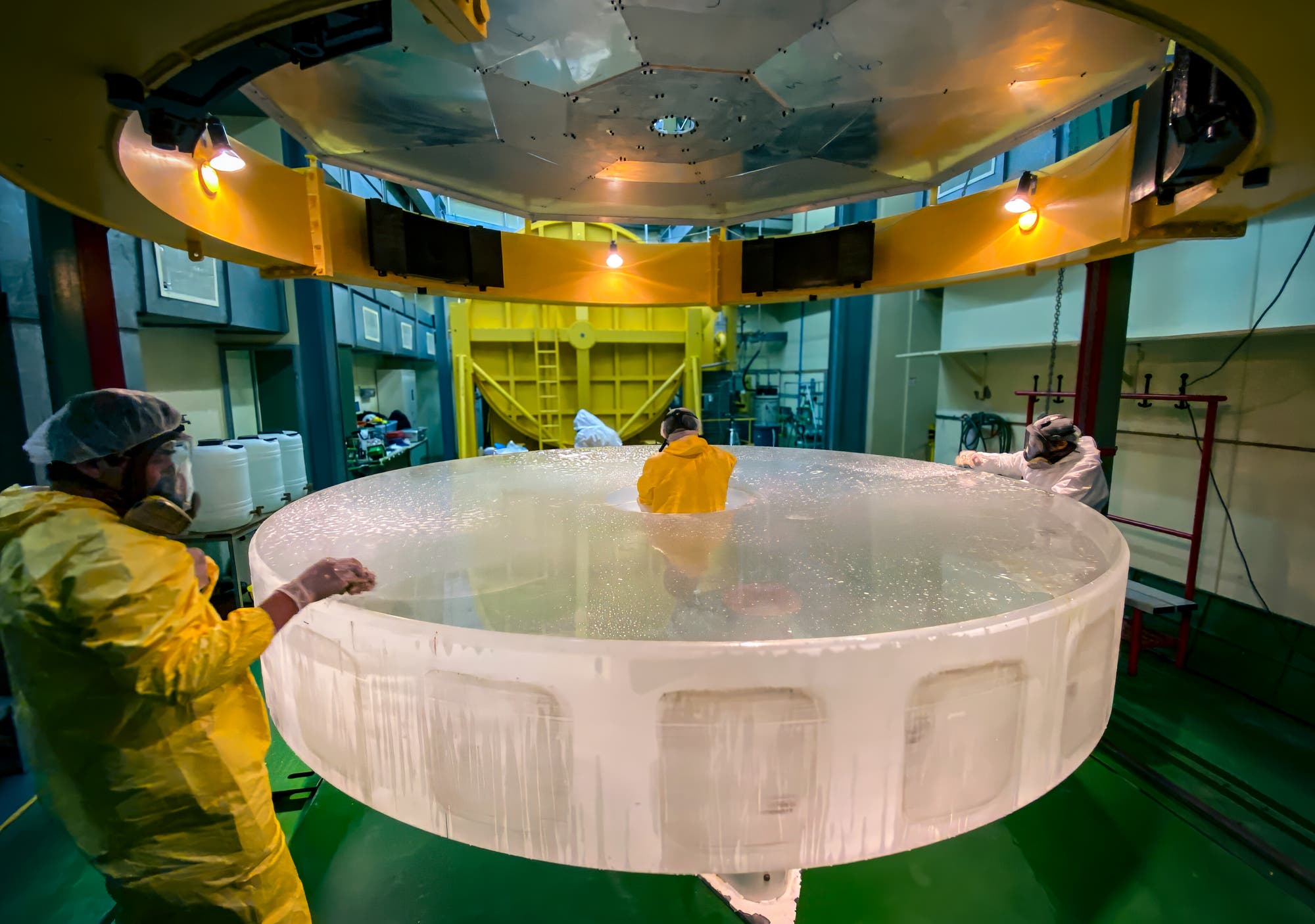 Drei Personen in Schutzkleidung und Atemmasken arbeiten in einem Labor an einem großen, runden, transparenten Objekt. Es ist der Hauptspiegel des 3,6-Meter-Teleskops der Europäischen Südsternwarte ESO in Chile, der hier gerade einer Reinigung unterzogen wird. Die Umgebung ist industriell, mit gelben und grünen Elementen. Oben hängt eine große, runde, gelbe Struktur mit Lichtern.