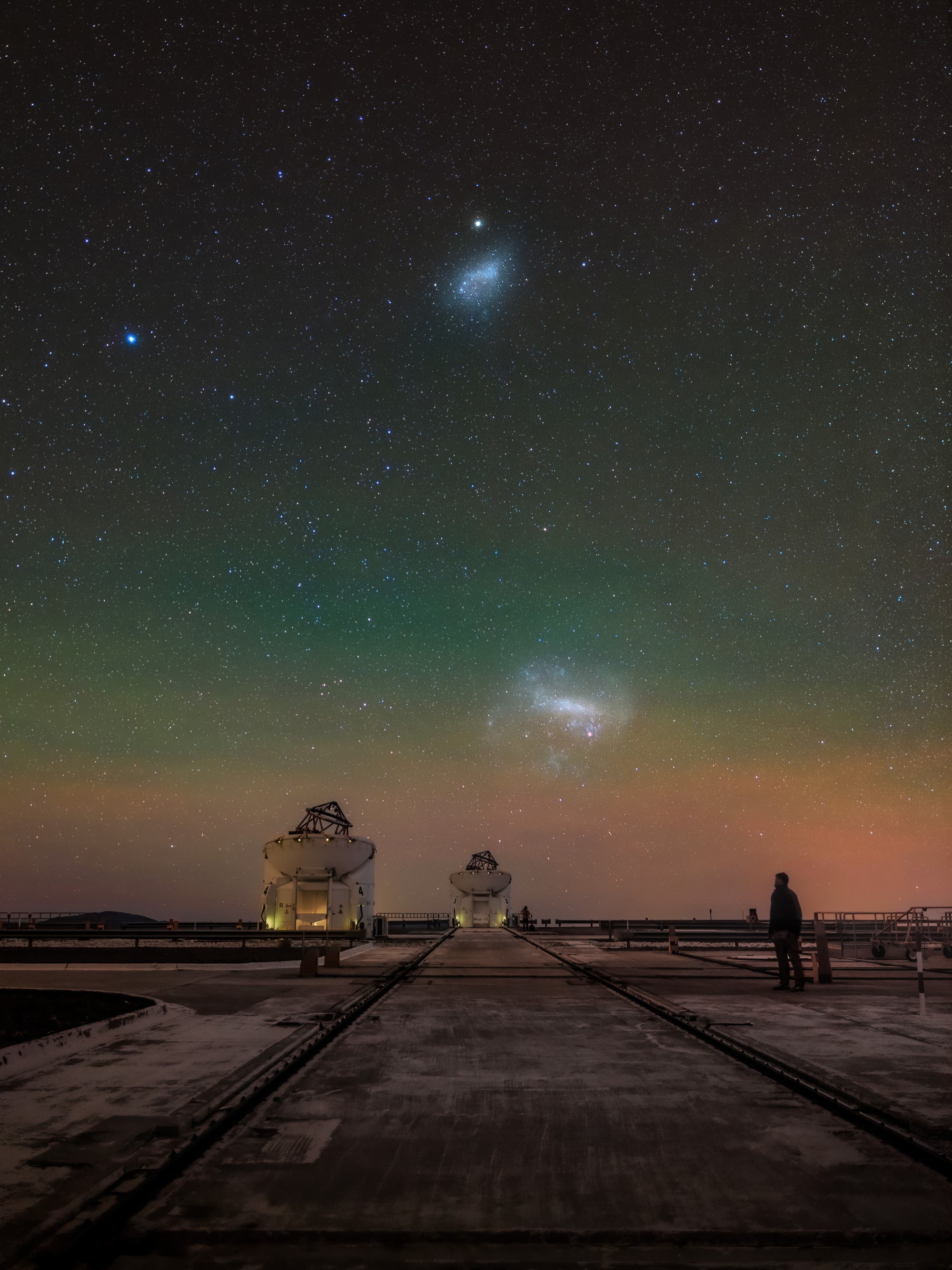 Ein nächtliches astronomisches Bild zeigt einen klaren Sternenhimmel mit zwei markanten Galaxien, der Großen und der Kleinen Magellanschen Wolke, die über einem Observatorium leuchten. Im Vordergrund sind mehrere Teleskope auf Schienen zu sehen, die auf einer Plattform stehen. Eine Person steht rechts und blickt in den Himmel. Der Himmel zeigt einen sanften Farbverlauf von Grün bis Orange, was auf atmosphärische Effekte hinweist.