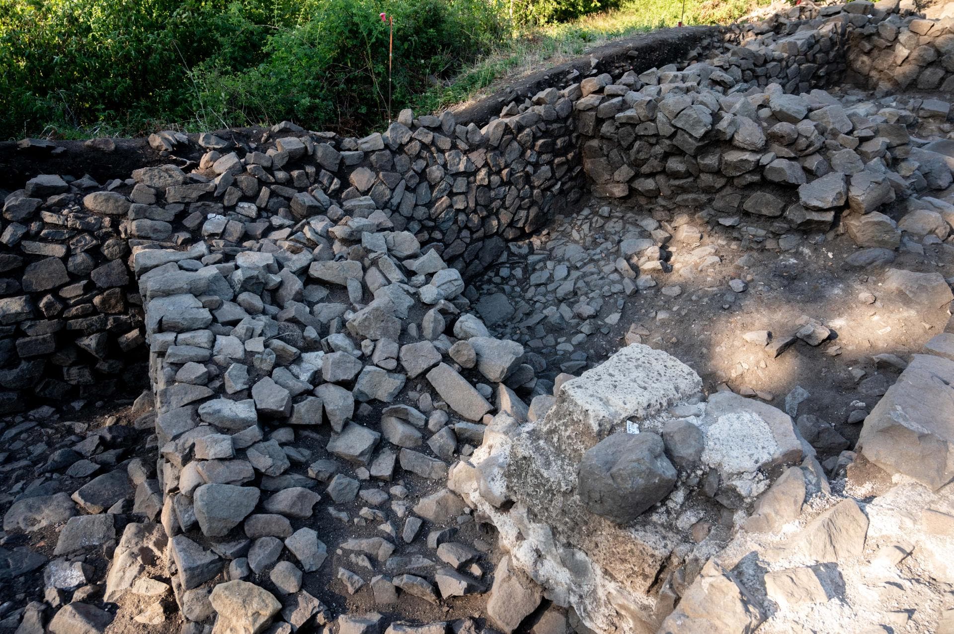 Eine archäologische Ausgrabungsstätte mit freigelegten Steinmauern und einem Boden aus unregelmäßig geformten Steinen. Die Mauern bestehen aus grob behauenen Steinen, die in mehreren Schichten übereinander gestapelt sind. Im Hintergrund ist Vegetation zu sehen, die die Ausgrabungsstelle umgibt. Die Szene ist von Sonnenlicht beleuchtet, das Schatten auf den Boden wirft.