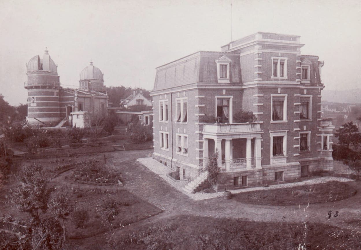 Ein historisches Foto zeigt ein großes, mehrstöckiges Backsteingebäude mit einem Mansardendach, umgeben von einem gepflegten Garten. Im Hintergrund sind zwei Kuppelgebäude zu sehen, die wie Observatorien aussehen. Der Garten ist mit Wegen und Pflanzen gestaltet. Die Szene vermittelt einen Eindruck von Ruhe und architektonischer Eleganz.