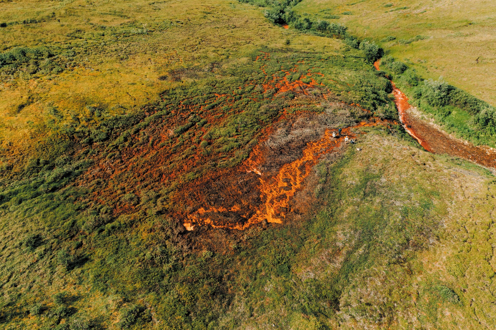 Luftaufnahme eines grün bewachsenen Landstrichs, der durch einen rotorange schimmernden Fluss zerschnitten wird. Die Erde um den Fluss herum ist tot und sieht aus wie verbrannt