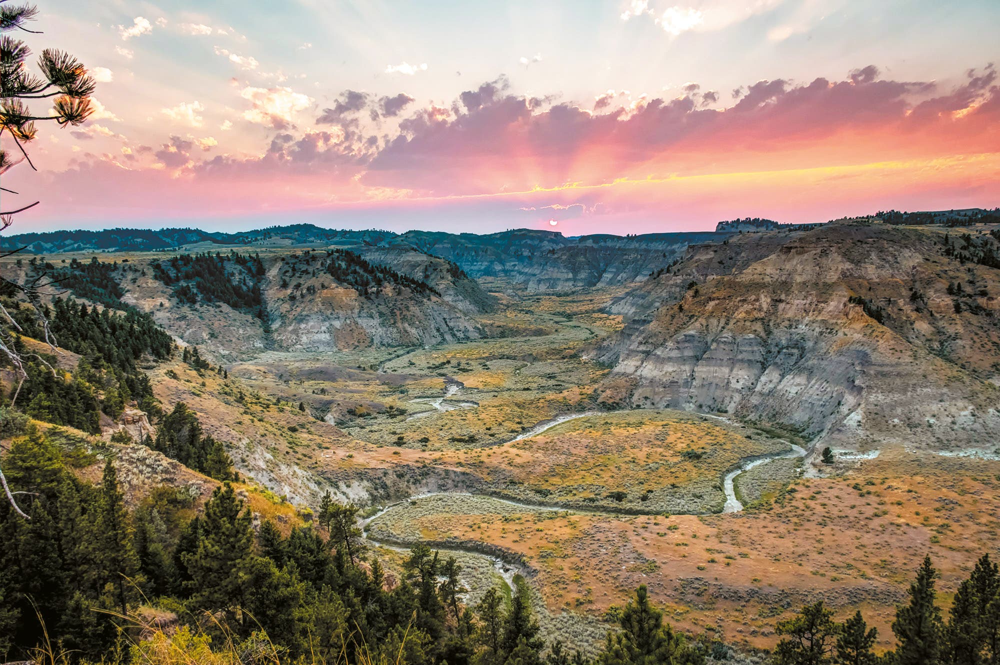 Weite Landschaft mit tiefen Schluchten und sanften Hügeln, die von einem Fluss durchzogen sind. Der Himmel zeigt einen dramatischen Sonnenuntergang mit rosa und orangefarbenen Wolken. Im Vordergrund sind dichte Wälder zu sehen. Die Szene vermittelt Ruhe und natürliche Schönheit.