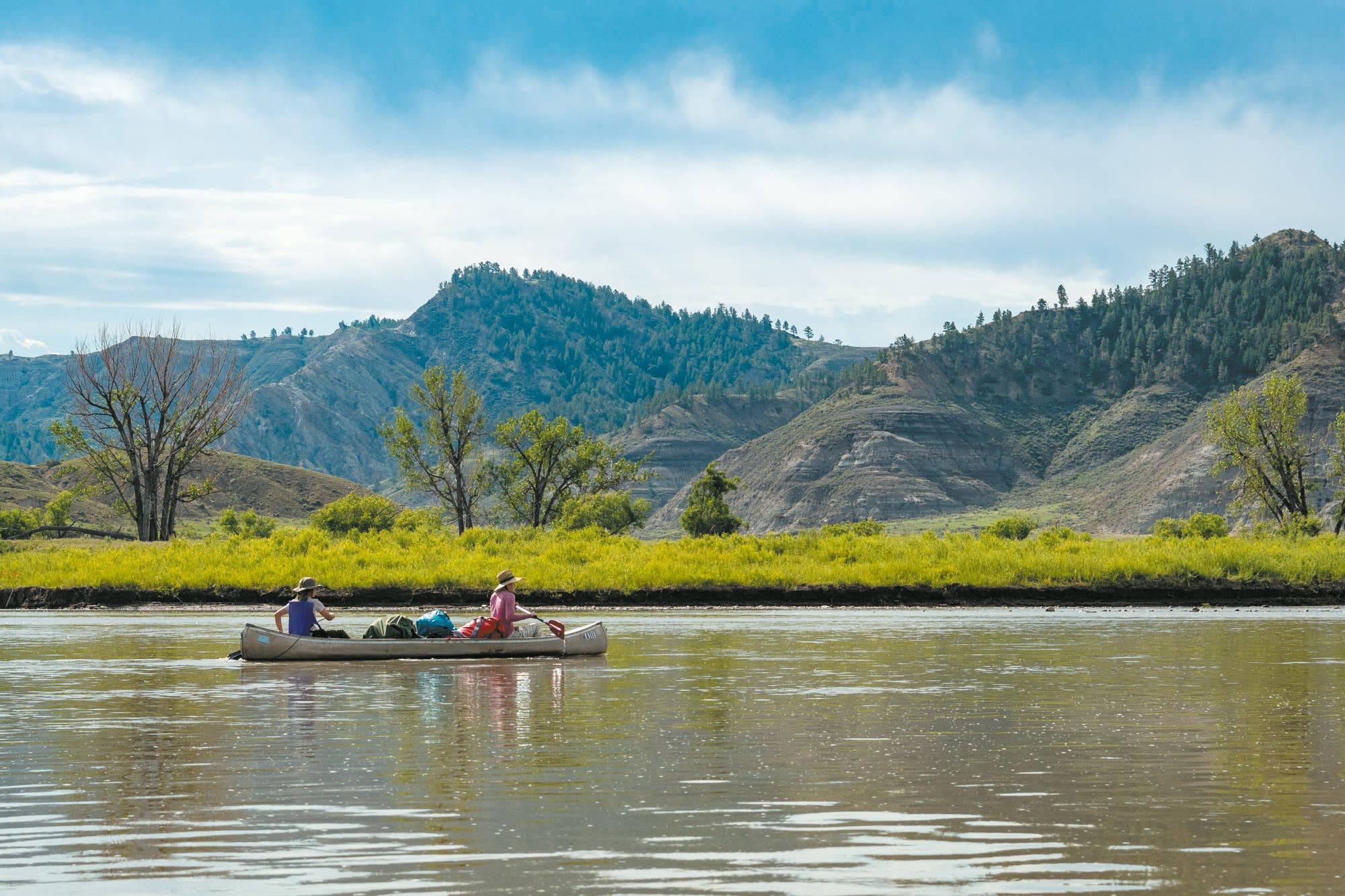 Zwei Personen paddeln in einem Kanu auf einem ruhigen Fluss, umgeben von einer malerischen Berglandschaft mit grünen Hügeln und Bäumen. Der Himmel ist leicht bewölkt. Die Szene vermittelt Ruhe und Naturverbundenheit.