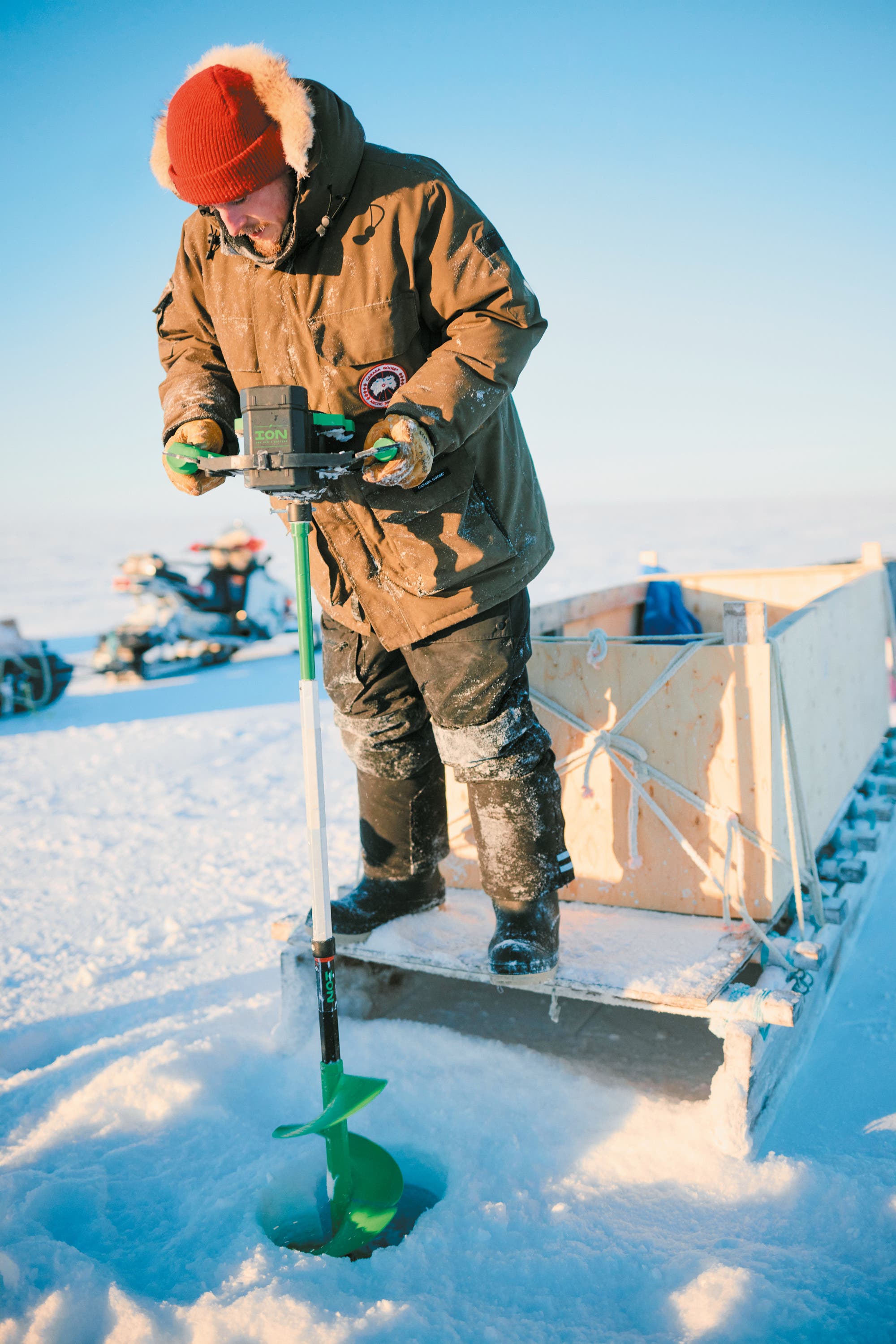 Eine Person in warmer Winterkleidung bohrt mit einem Eisbohrer ein Loch in eine gefrorene Oberfläche. Im Hintergrund sind ein Schlitten und ein Schneemobil zu sehen. Die Szene findet in einer verschneiten, offenen Landschaft bei klarem Himmel statt.