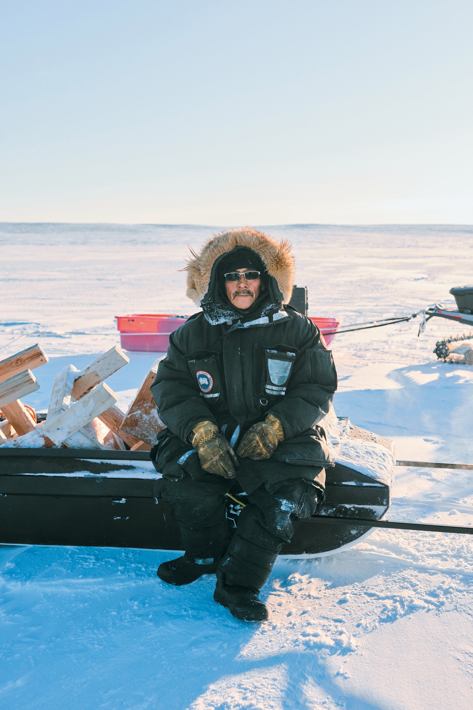 Eine Person in warmer Winterkleidung sitzt auf einem Schlitten in einer verschneiten Landschaft. Der Schlitten ist mit Holz beladen. Die Person trägt eine dicke Jacke mit Fellkapuze, Handschuhe und eine Sonnenbrille. Im Hintergrund erstreckt sich eine weite, schneebedeckte Ebene unter einem klaren Himmel.