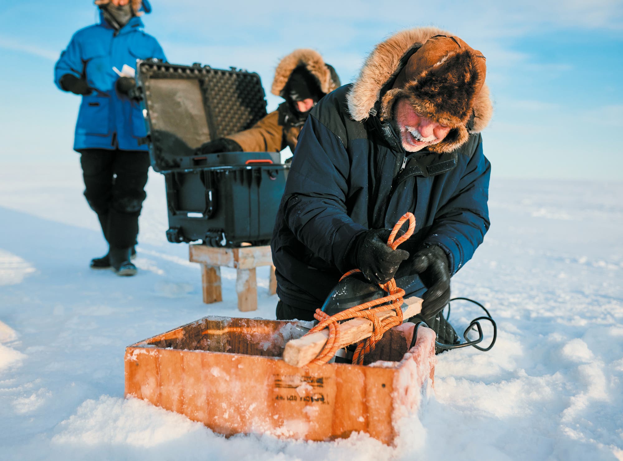 Eine Gruppe von Forschern arbeitet in einer verschneiten, arktischen Umgebung. Im Vordergrund kniet eine Person in einem dicken, pelzbesetzten Mantel und bearbeitet ein hölzernes Gerät in einer Kiste. Im Hintergrund stehen zwei weitere Personen, eine öffnet einen großen schwarzen Koffer. Der Himmel ist klar und blau. Die Szene vermittelt eine wissenschaftliche Expedition in einer kalten Region.