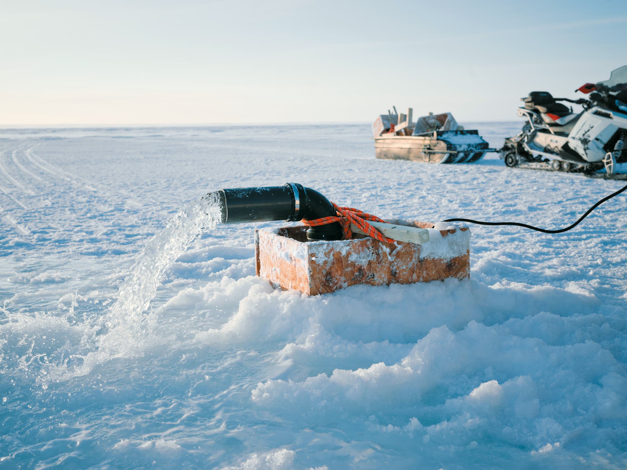 Ein schwarzes Rohr, das auf einem schneebedeckten Block montiert ist, pumpt Wasser in eine gefrorene Landschaft. Im Hintergrund sind ein Schlitten mit Ausrüstung und ein Schneemobil zu sehen. Die Szene zeigt eine winterliche Umgebung mit klarem Himmel.