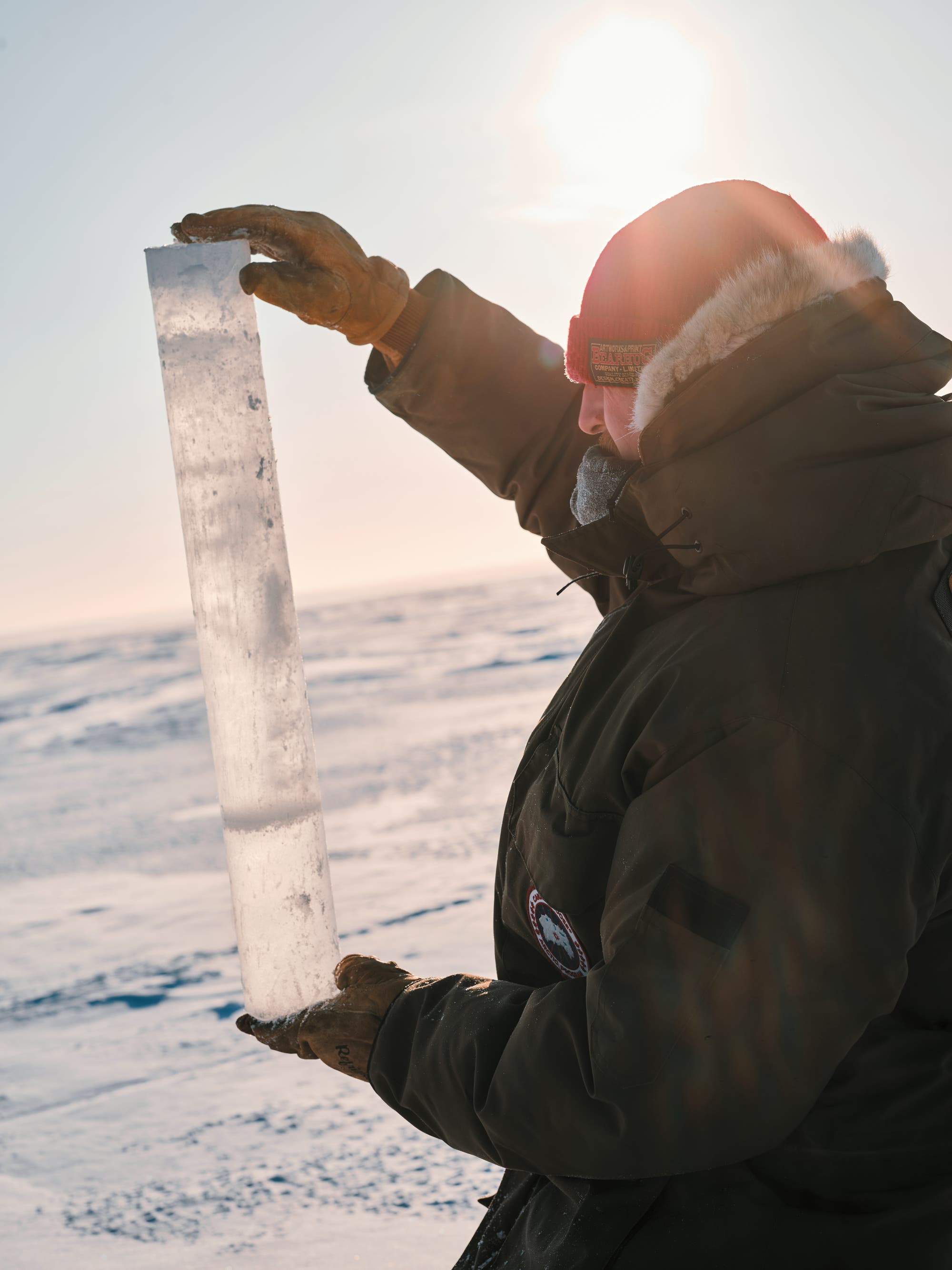 Eine Person in warmer Winterkleidung hält einen langen, transparenten Eisbohrkern gegen das Licht. Der Hintergrund zeigt eine verschneite, weite Landschaft unter einem klaren Himmel mit der Sonne im Hintergrund. Die Szene vermittelt eine wissenschaftliche Untersuchung in einer kalten Umgebung.