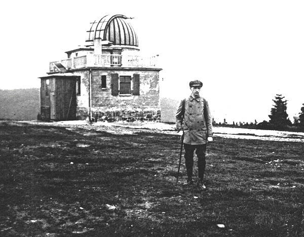 Ein Mann steht auf einer Wiese vor einem kleinen Observatorium mit einer Kuppel. Der Himmel ist bewölkt, und im Hintergrund sind Bäume zu sehen. Der Mann trägt einen Mantel und eine Mütze und hält einen Stock. Das Gebäude hat Fensterläden, eine Tür an der Seite und oben ein Geländer.
