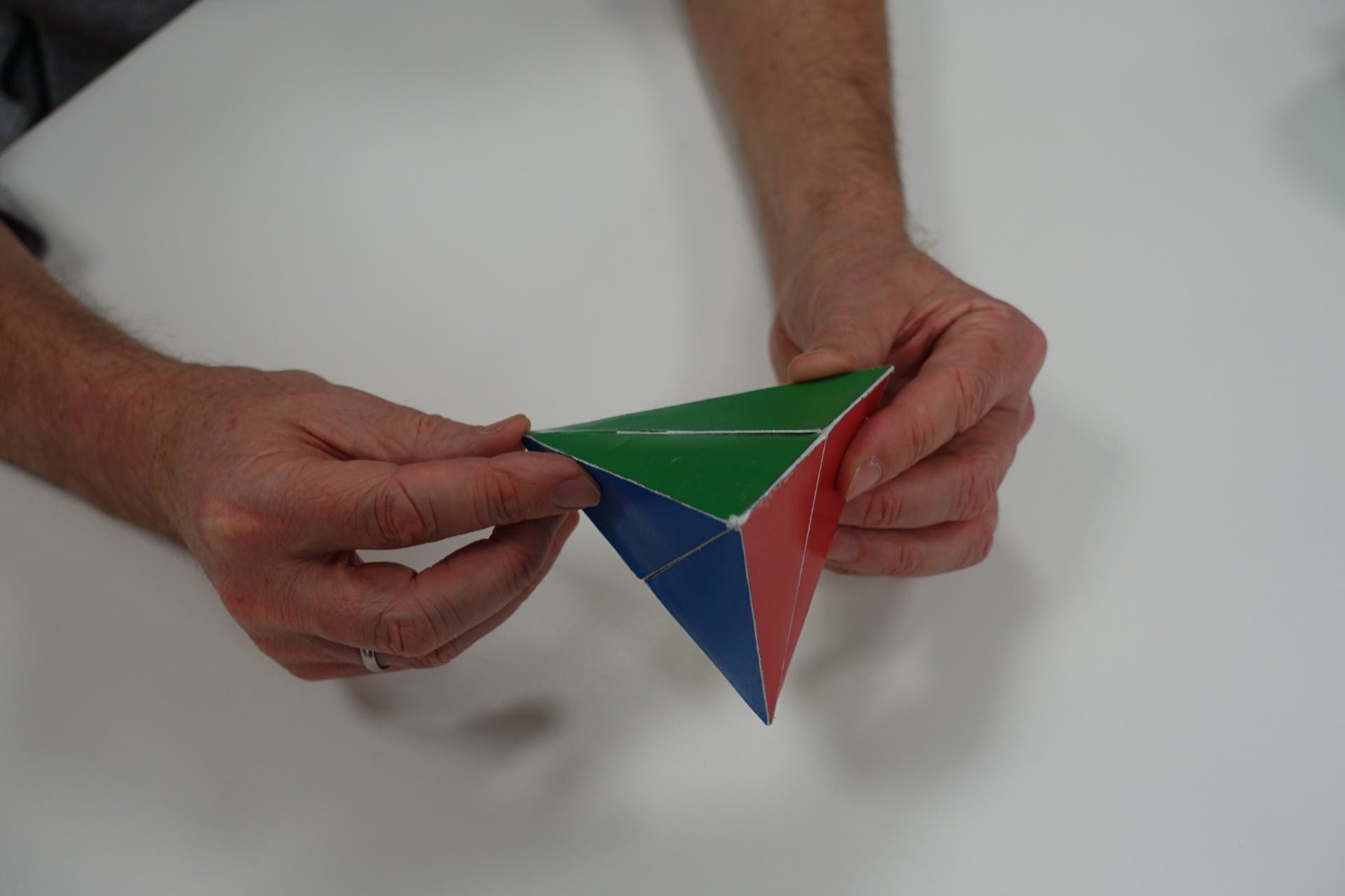 Eine Person hält ein Origami-Modell in den Händen. Das Modell hat eine geometrische Form mit blauen und weißen Flächen. Die Hände der Person sind auf einem weißen Tisch sichtbar.