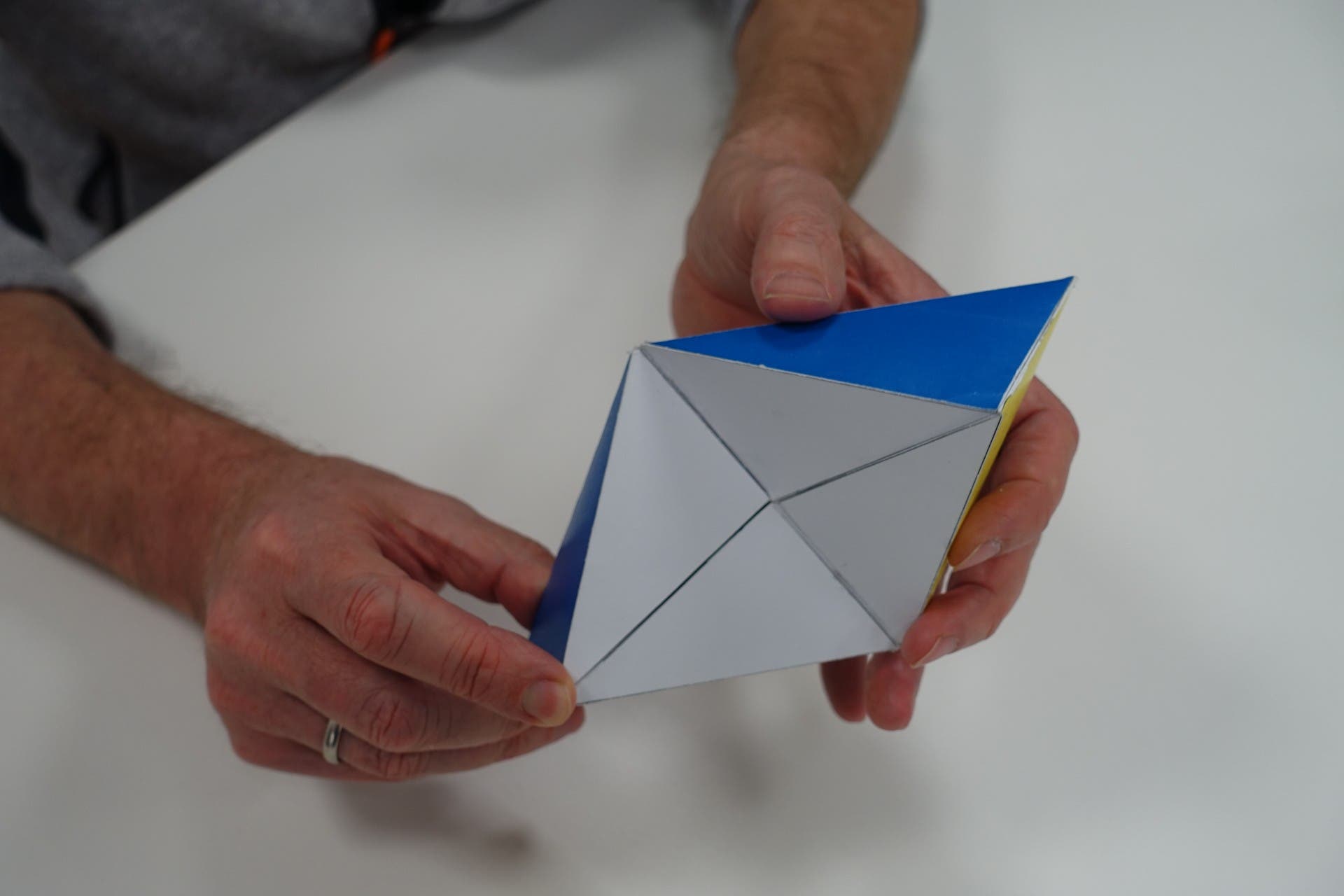 Eine Person hält ein Origami-Modell in Form eines dreidimensionalen geometrischen Körpers. Das Modell hat blaue und weiße Flächen und ist aus Papier gefaltet. Die Hände der Person sind sichtbar, während sie das Modell über einem weißen Tisch hält.