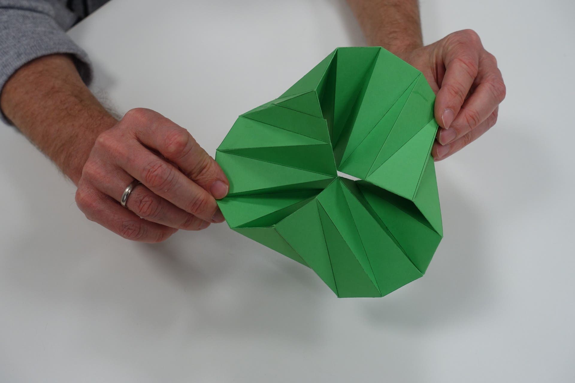 Zwei Hände halten ein komplex gefaltetes Origami-Modell aus grünem Papier, das eine spiralförmige, dreidimensionale Struktur bildet. Die Hände gehören einer Person mit einem grauen Pullover, die einen silbernen Ring am linken Ringfinger trägt. Der Hintergrund ist ein weißer Tisch.