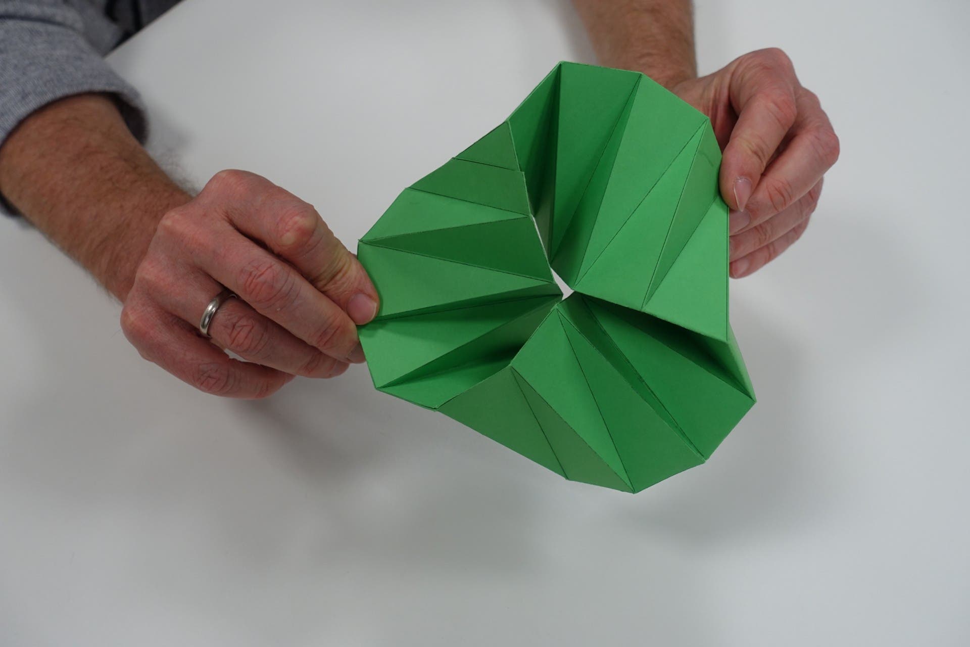 Eine Person hält ein grünes, gefaltetes Origami-Modell in den Händen. Das Modell hat eine komplexe, spiralförmige Struktur. Die Hände sind leicht gebräunt, und an einem Finger ist ein silberner Ring zu sehen. Der Hintergrund ist ein weißer Tisch.