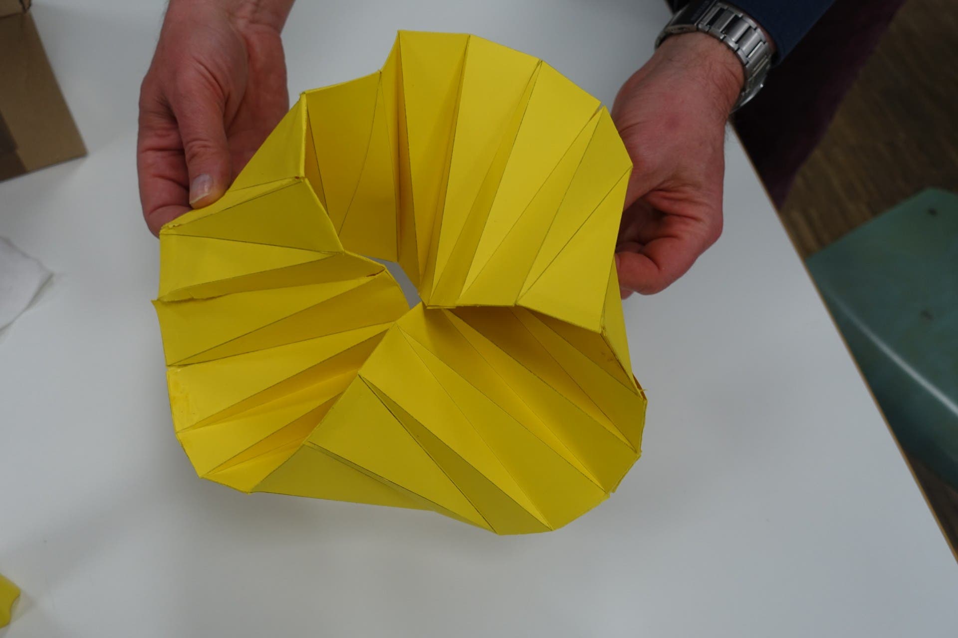 Zwei Hände halten ein gelbes, spiralförmiges Origami-Modell aus Papier über einem weißen Tisch. Das Modell besteht aus gefalteten, geometrischen Segmenten, die eine komplexe, dreidimensionale Form bilden.