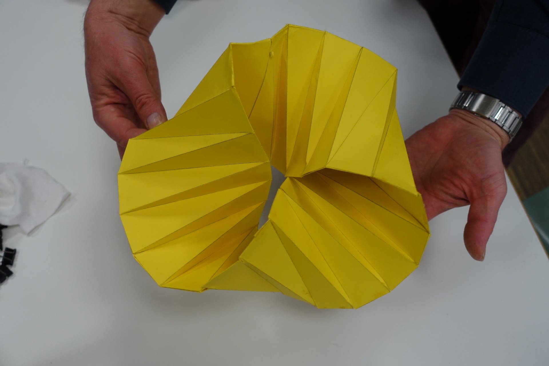 Zwei Hände halten ein gelbes, geometrisch gefaltetes Papierobjekt auf einem weißen Tisch. Das Objekt hat eine komplexe, spiralförmige Struktur mit klaren, gefalteten Kanten. Im Hintergrund sind unscharf weiße und schwarze Gegenstände zu erkennen.