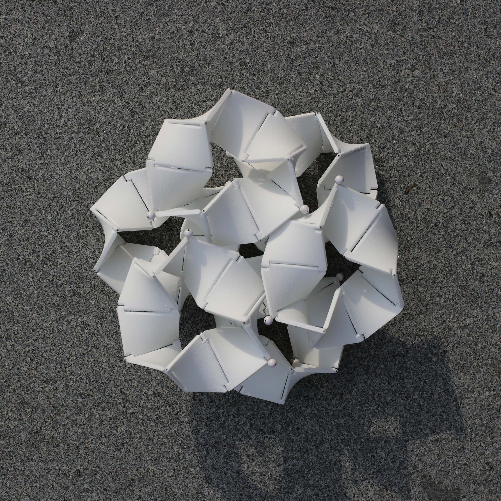 Eine komplexe, geometrische Struktur aus weißen, miteinander verbundenen Modulen liegt auf einer grauen, körnigen Oberfläche. Die Struktur besteht aus mehreren sechseckigen Formen, die durch kleine Kugelgelenke verbunden sind, und wirft einen Schatten auf den Untergrund.
