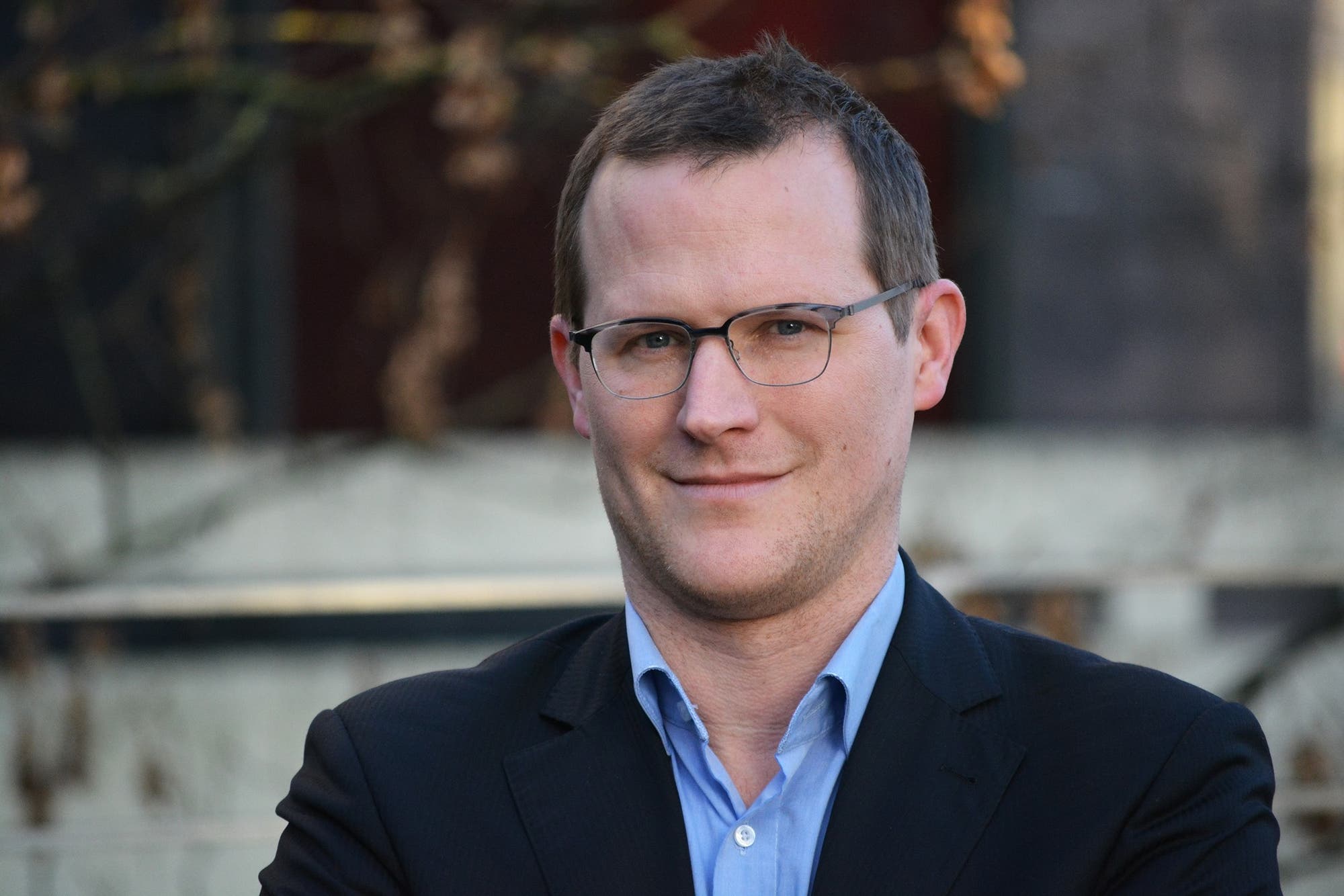 Ein Mann mit kurzen braunen Haaren, Brille, blauem Hemd und dunklem Jacket lächelt in die Kamera. Der Bildhintergrund ist verschwommen