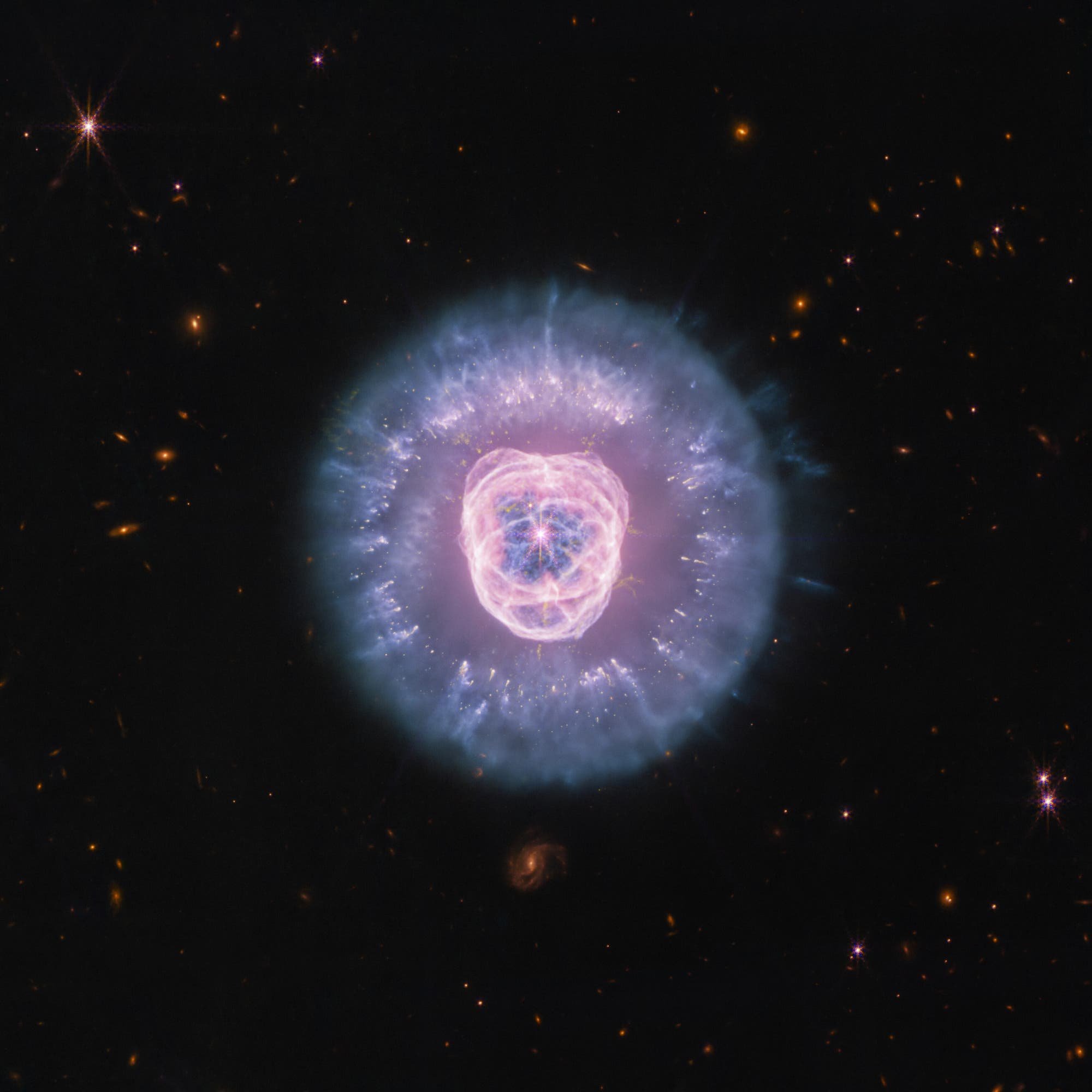 Ein farbenfrohes astronomisches Bild zeigt einen planetarischen Nebel im Weltraum. Im Zentrum befindet sich eine komplexe, symmetrische Struktur aus leuchtenden Gasen in Rosa- und Blaut&ouml;nen, die von einem &auml;u&szlig;eren, bl&auml;ulichen Halo umgeben ist. Der Hintergrund ist mit zahlreichen kleinen, leuchtenden Sternen und Galaxien &uuml;bers&auml;t. Der Nebel hebt sich deutlich vom dunklen, sternenreichen Hintergrund ab und zeigt die dynamische Sch&ouml;nheit kosmischer Ph&auml;nomene.