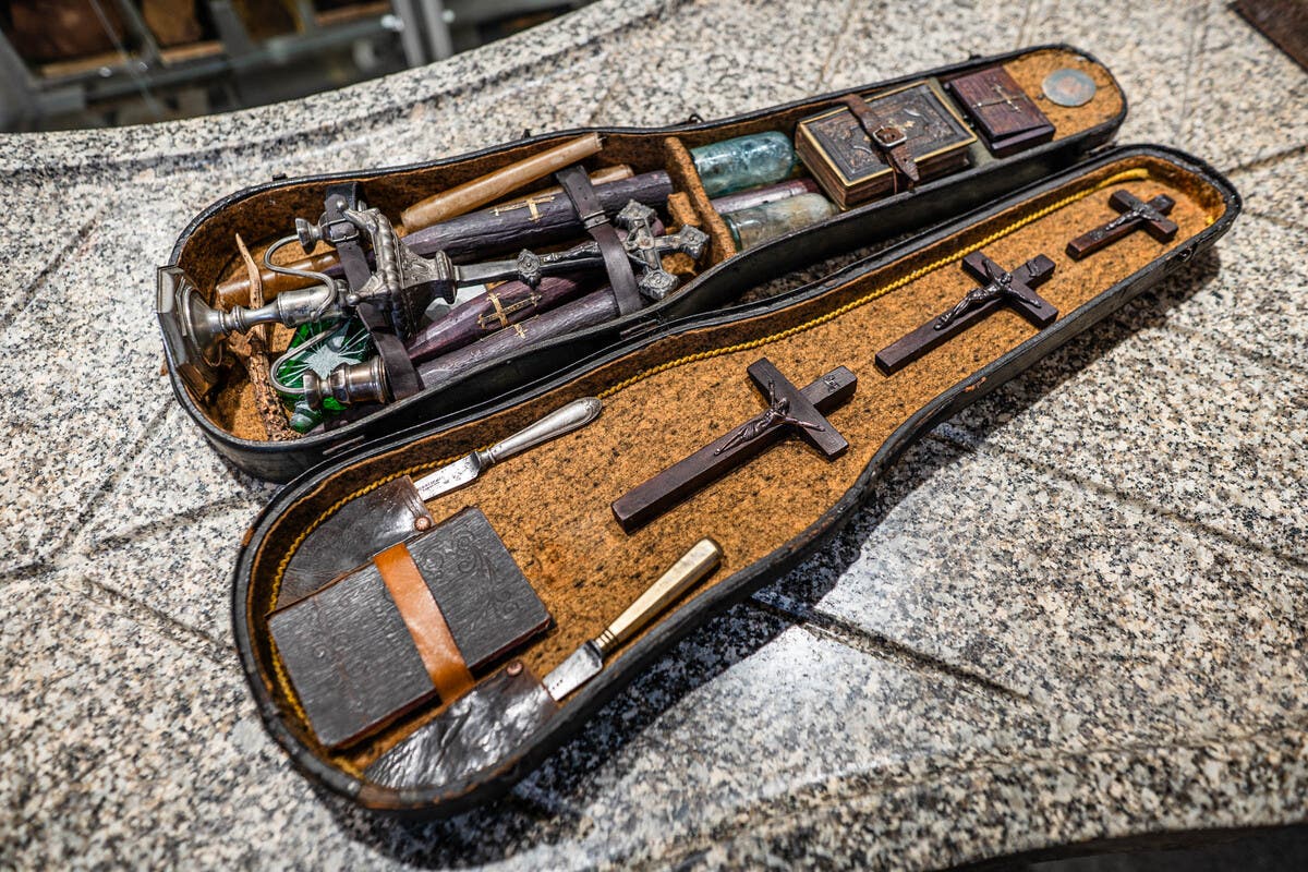 Ein offener Koffer auf einer Granitoberfläche zeigt eine Sammlung von Gegenständen, darunter mehrere Kreuze, Flaschen, ein Buch und Messer. Die Gegenstände sind sorgfältig in einem mit Stoff ausgekleideten Innenraum angeordnet. Der Koffer scheint antik zu sein und die Anordnung der Objekte deutet auf ein historisches oder rituelles Set hin. Ein offener Koffer auf einer Granitoberfläche zeigt eine Sammlung von Gegenständen, darunter mehrere Kreuze, Flaschen, ein Buch und Messer. Die Gegenstände sind sorgfältig in einem mit Stoff ausgekleideten Innenraum angeordnet. Der Koffer scheint antik zu sein und die Anordnung der Objekte deutet auf ein historisches oder rituelles Set hin.