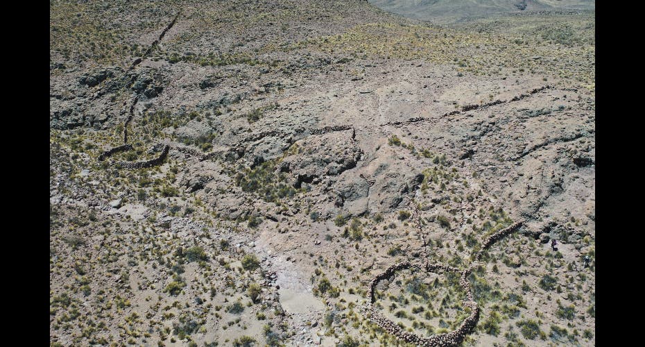 Luftaufnahme einer trockenen, felsigen Landschaft mit spärlicher Vegetation. Im Bild sind mehrere kurvenreiche, niedrige Steinmauern zu sehen, die sich durch das Gelände schlängeln. Die Mauern bilden unregelmäßige Muster und heben sich von der natürlichen Umgebung ab. Die Szene vermittelt einen Eindruck von Abgeschiedenheit und natürlicher Schönheit.