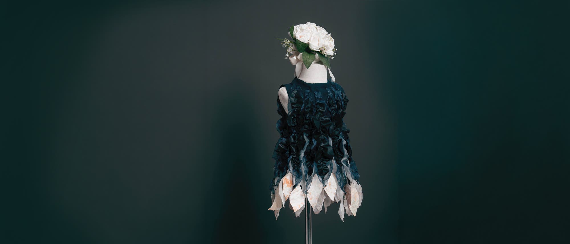 Eine Schaufensterpuppe tr&auml;gt ein kunstvolles Kleid mit dunklen, gerafften Stofflagen und einem Saum aus blatt&auml;hnlichen, wei&szlig;en Elementen. Auf dem Kopf der Puppe befindet sich ein Kranz aus wei&szlig;en Rosen. Der Hintergrund ist dunkel und schlicht, wodurch das Kleid im Fokus steht.