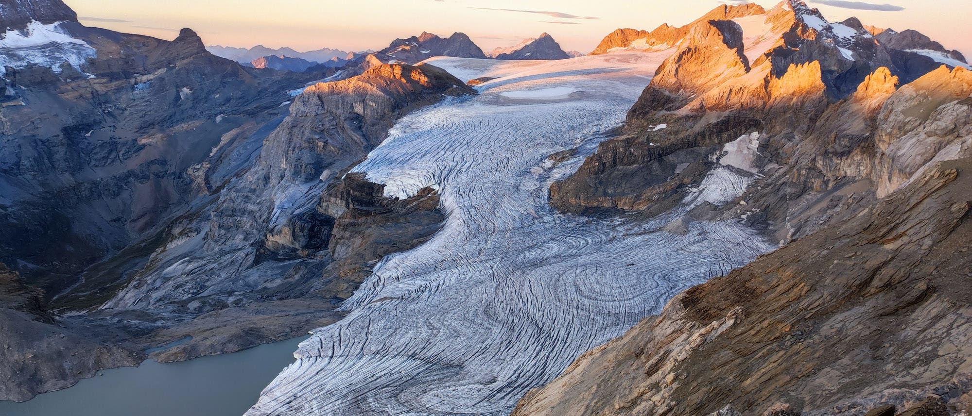 Blick auf einen Gletscher in einer Berglandschaft bei Sonnenuntergang. Der Gletscher zieht sich durch ein Tal, umgeben von felsigen Bergen, die von der untergehenden Sonne in warmes Licht getaucht werden. Im Vordergrund sind Ger&ouml;ll und Felsen zu sehen, w&auml;hrend im Hintergrund schneebedeckte Gipfel den Horizont s&auml;umen. Der Himmel ist klar mit wenigen Wolken.
