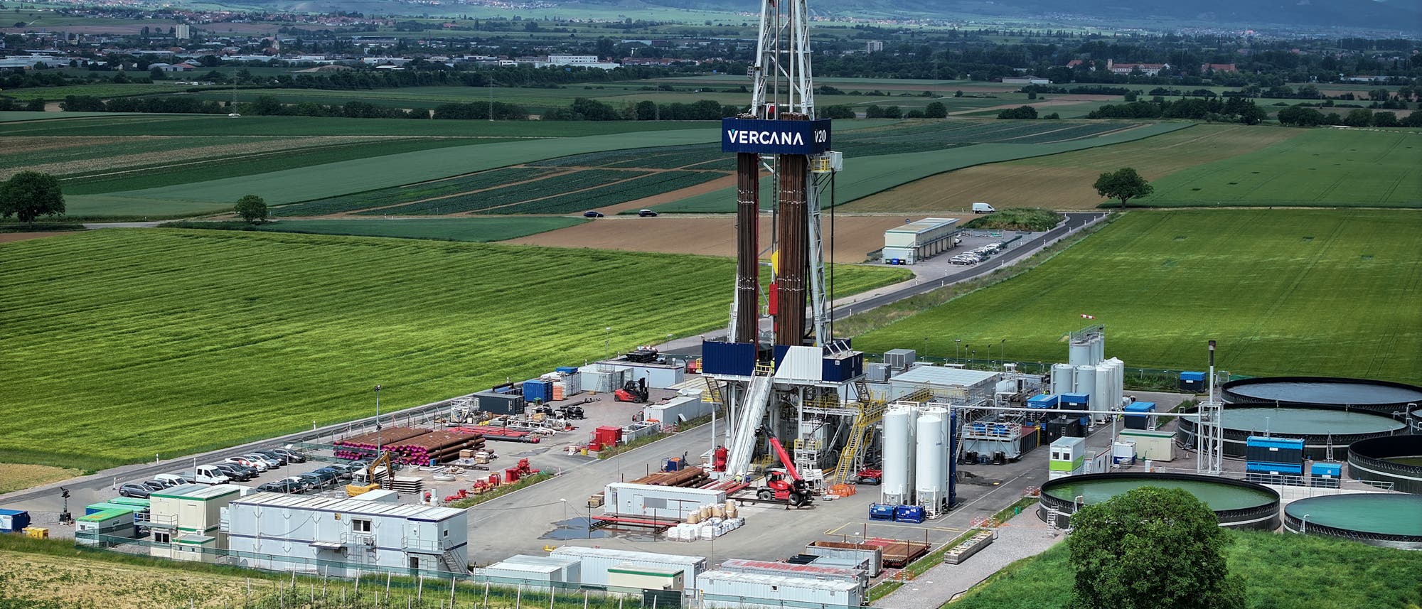 Eine Bohrplattform inmitten einer weitläufigen, grünen Landschaft mit Feldern und Hügeln im Hintergrund. Die Plattform ist von verschiedenen Containern und Tanks umgeben. Auf der Plattform ist der Schriftzug "VERCANA" zu sehen. Die Szene zeigt eine industrielle Anlage in einer ländlichen Umgebung unter einem bewölkten Himmel.