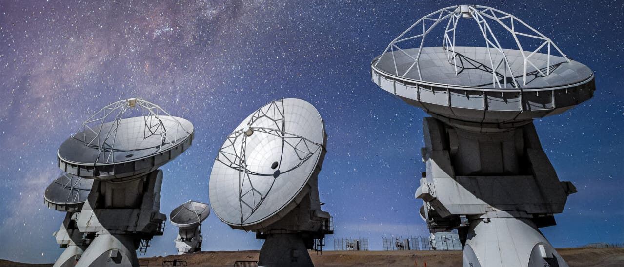 Eine Gruppe von großen Radioteleskopen steht auf einer trockenen, erdigen Fläche unter einem klaren Nachthimmel, der mit unzähligen Sternen übersät ist. Im Hintergrund ist die Milchstraße als leuchtendes Band sichtbar. Die Teleskopschüsseln sind in verschiedene Richtungen ausgerichtet und scheinen den Himmel zu beobachten.