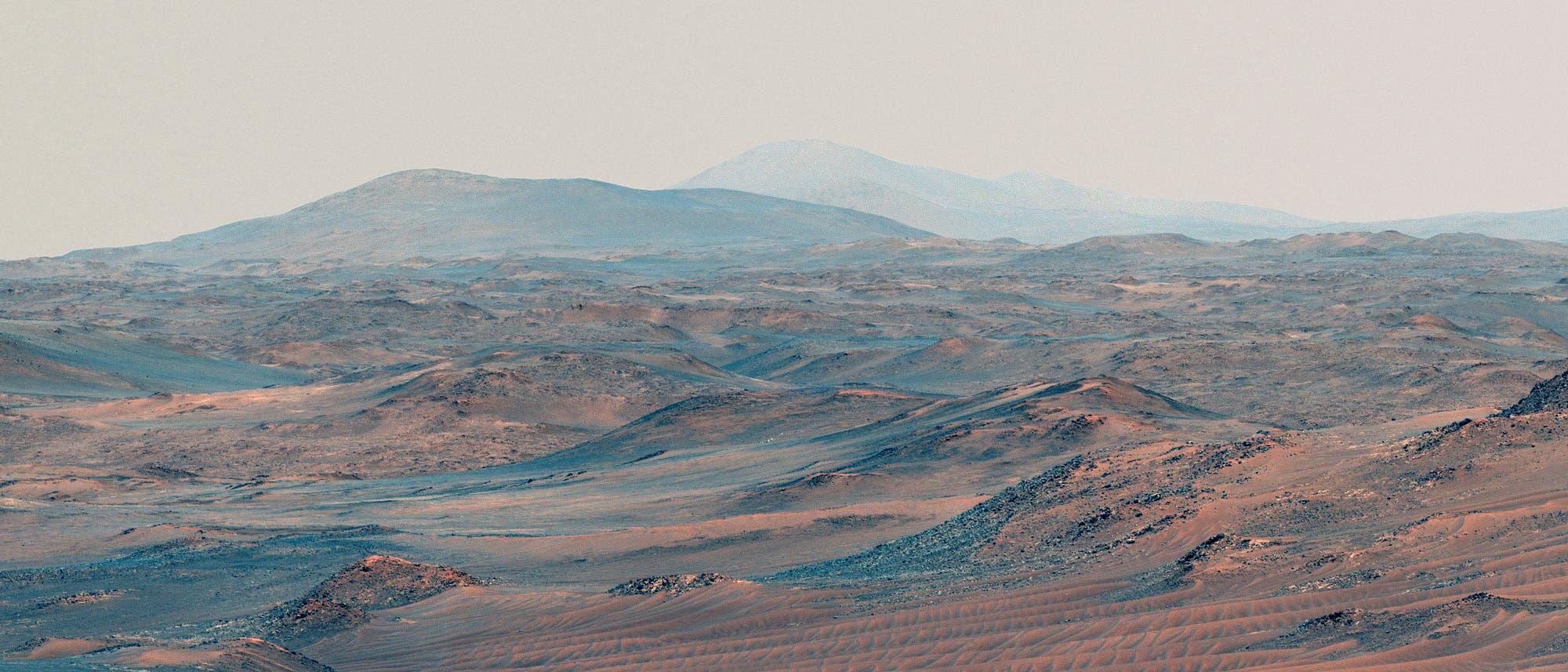 Weite, karge W&uuml;stenlandschaft mit sanften H&uuml;geln und Bergen im Hintergrund. Die Oberfl&auml;che ist von sandigen und felsigen Strukturen gepr&auml;gt, die in warmen Erdt&ouml;nen leuchten. Der Himmel ist klar und hell, was die Weite der Landschaft betont.