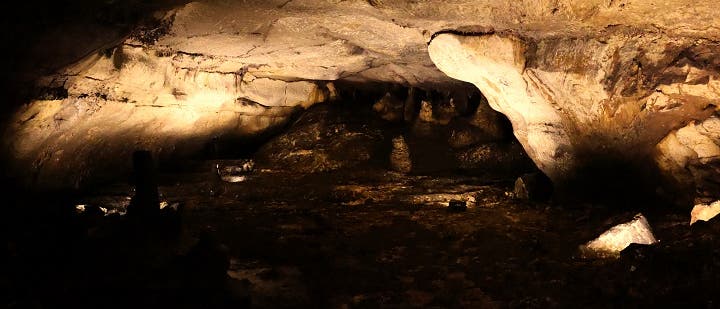 Eine beleuchtete H&ouml;hle mit unregelm&auml;&szlig;igen Felsformationen und einer niedrigen Decke. Der Boden ist uneben und es gibt einige hervorstehende Steine. Die Beleuchtung hebt die Textur der Felsw&auml;nde hervor und erzeugt Schatten, die die Tiefe der H&ouml;hle betonen.