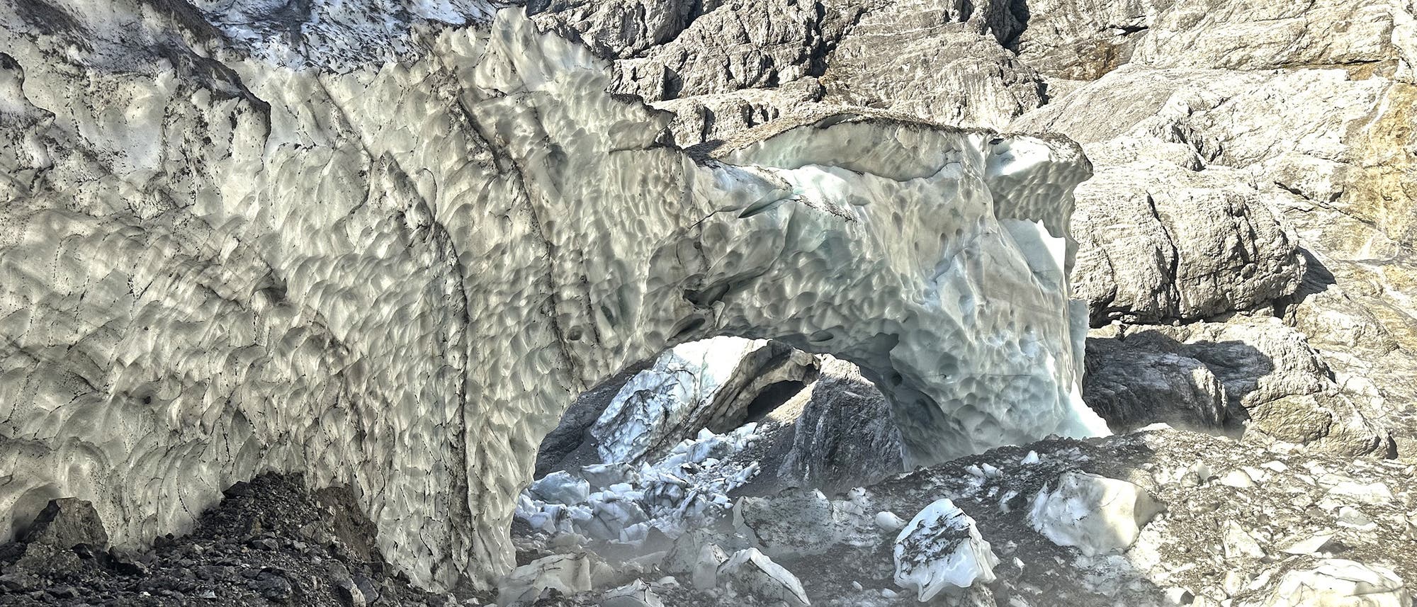 Eine natürliche Eishöhle in einem Gletscher, umgeben von felsigem Gelände. Die Höhle zeigt eine gewölbte Struktur mit einer unregelmäßigen, texturierten Oberfläche aus Eis. Im Hintergrund sind steinige Felsformationen sichtbar. Die Szene vermittelt eine kühle, raue Umgebung in einer alpinen Landschaft.