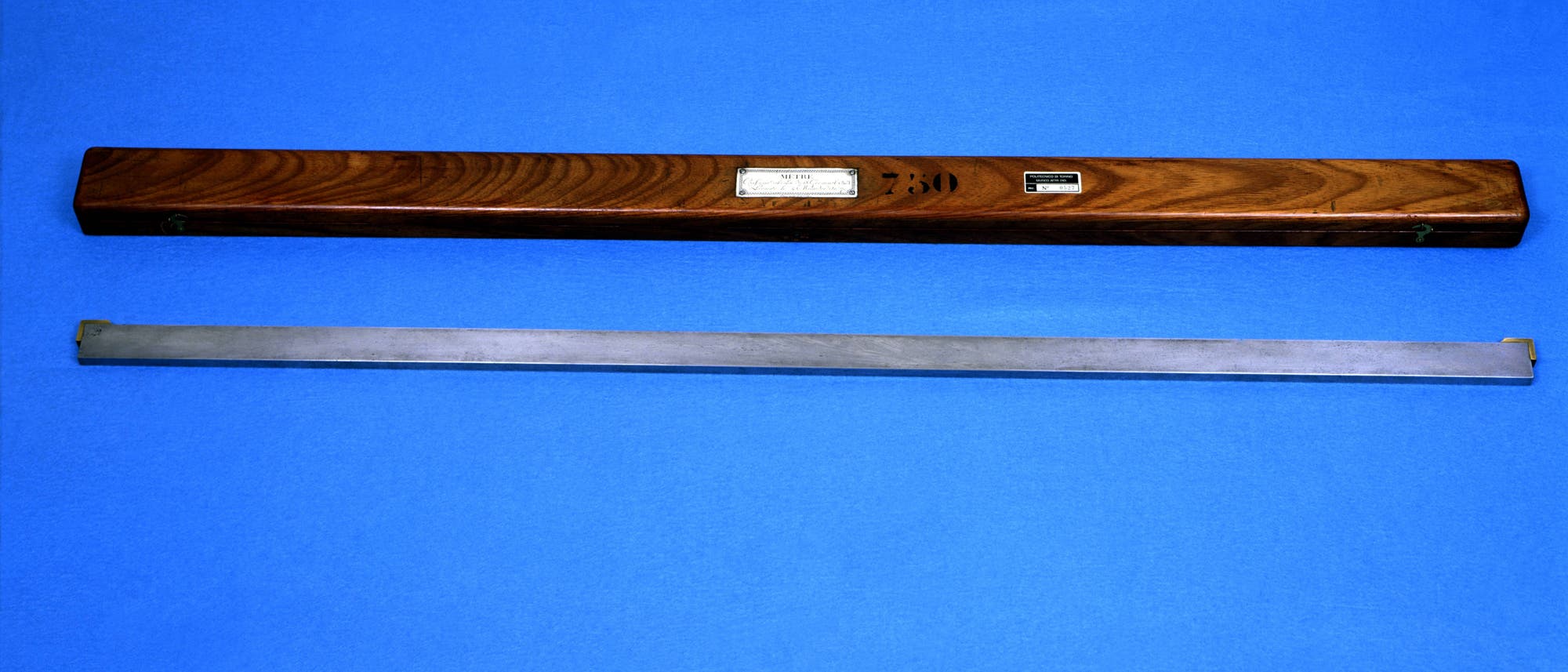 Ein hölzernes Etui mit Maserung und zwei Etiketten liegt auf einem blauen Hintergrund. Darunter befindet sich ein langer, schmaler Metallstab. Das Etui trägt die Aufschrift "730". Ein hölzernes Etui mit Maserung und zwei Etiketten liegt auf einem blauen Hintergrund. Darunter befindet sich ein langer, schmaler Metallstab. Das Etui trägt die Aufschrift "730".