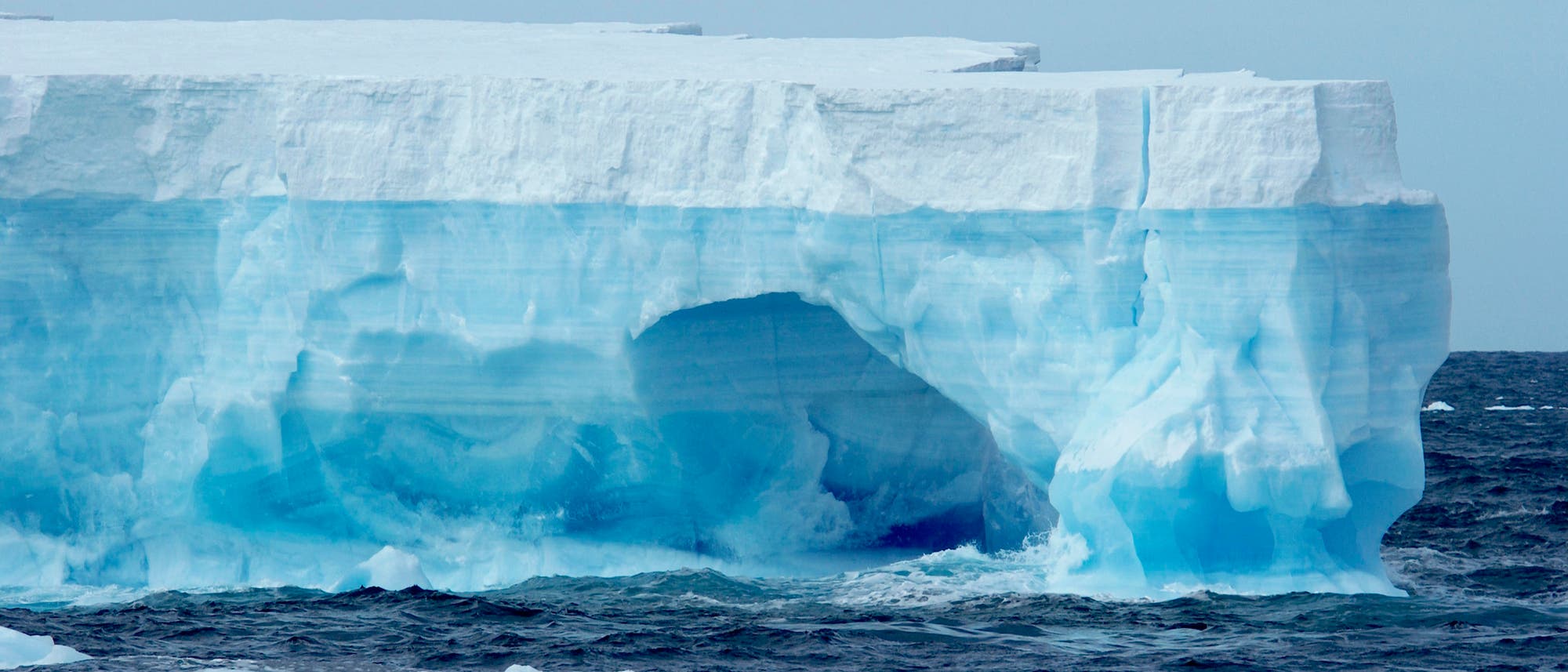 Ein blauweißer Tafeleisberg schwimmt durch unruhiges dunkelblaues Wasser im Südpolarmeer, kleinere Eisbrocken sind im Vordergrund, der Himmel ist milchig-blau