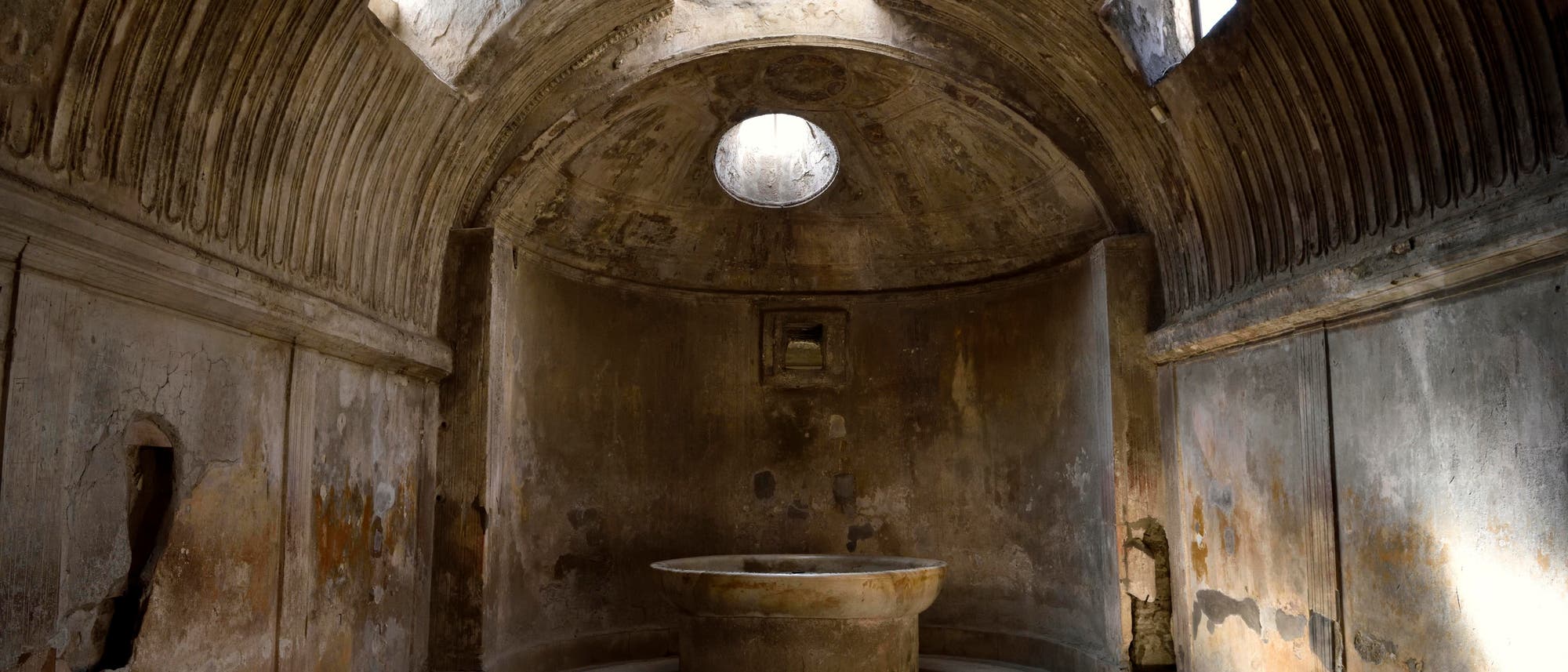 Ein antiker r&ouml;mischer Raum mit gew&ouml;lbter Decke und Licht&ouml;ffnungen, die nat&uuml;rliches Licht hereinlassen. In der Mitte steht ein gro&szlig;er, runder Steinbrunnen. Die W&auml;nde sind mit verblassten Fresken und Rissen versehen, die auf das Alter des Bauwerks hinweisen. Der Boden ist mit Mosaiken bedeckt.