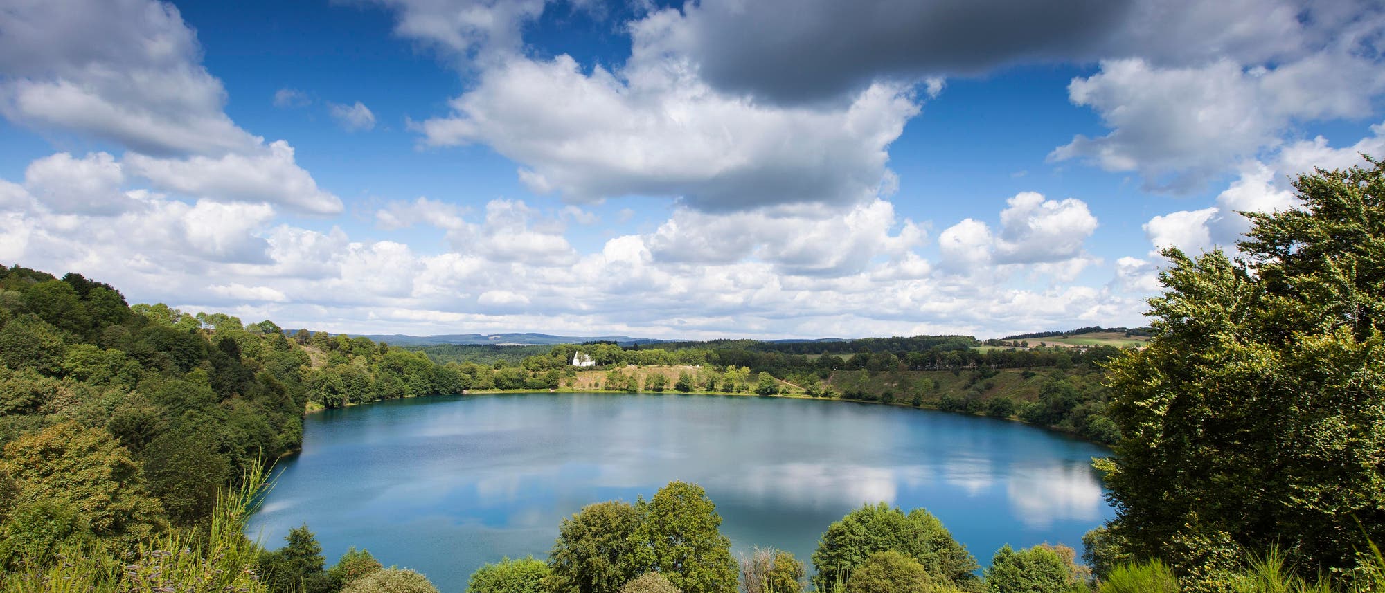Ein malerischer See, umgeben von üppigen, grünen Bäumen und sanften Hügeln unter einem blauen Himmel mit weißen Wolken. Die Landschaft strahlt Ruhe und natürliche Schönheit aus. Im Hintergrund sind entfernte Hügel zu sehen. Ein malerischer See, umgeben von üppigen, grünen Bäumen und sanften Hügeln unter einem blauen Himmel mit weißen Wolken. Die Landschaft strahlt Ruhe und natürliche Schönheit aus. Im Hintergrund sind entfernte Hügel zu sehen.