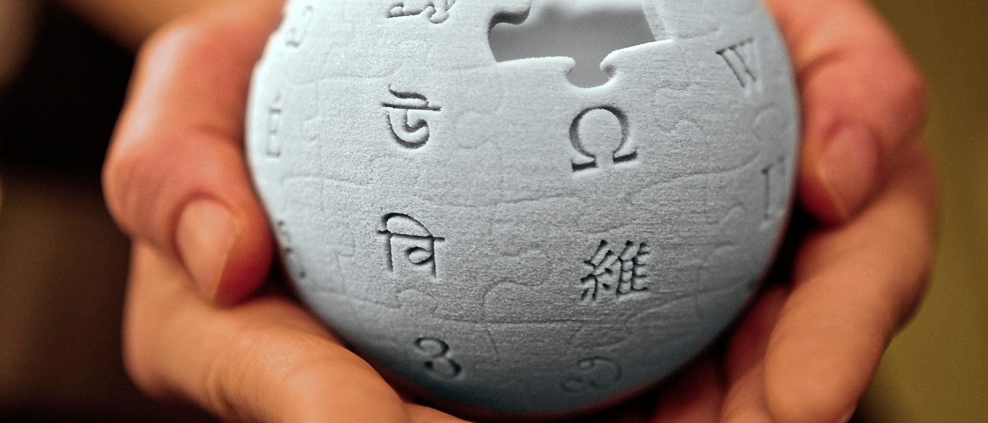 Zwei H&auml;nde halten eine dreidimensionale Nachbildung des Wikipedia-Logos, das aus Puzzleteilen besteht. Auf der Kugel sind verschiedene Schriftzeichen und Buchstaben aus verschiedenen Alphabeten eingraviert, darunter das griechische Omega und andere Symbole. Die Szene symbolisiert den globalen und kollaborativen Charakter von Wikipedia.