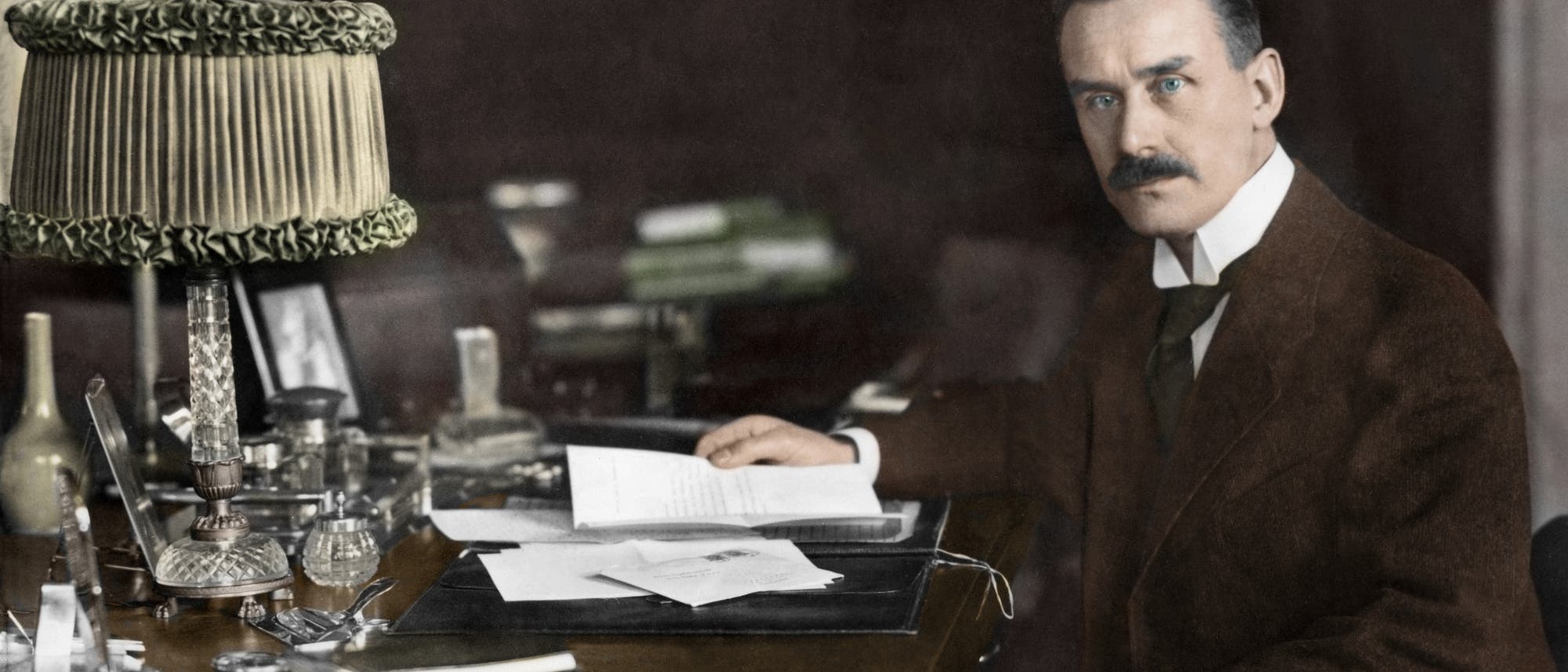 Ein Mann im Anzug sitzt an einem Schreibtisch, umgeben von Büchern und Papieren. Er hält ein Schriftstück in der rechten Hand und schaut nach links direkt in die Kamera. Auf dem Schreibtisch stehen eine verzierte Lampe, Schreibutensilien und ein Tintenfass. Im Hintergrund sind Bücherregale zu sehen. Die Atmosphäre ist ernst und erhaben. Ein Mann im Anzug sitzt an einem Schreibtisch, umgeben von Büchern und Papieren. Er hält ein Schriftstück in der rechten Hand und schaut nach links direkt in die Kamera. Auf dem Schreibtisch stehen eine verzierte Lampe, Schreibutensilien und ein Tintenfass. Im Hintergrund sind Bücherregale zu sehen. Die Atmosphäre ist ernst und erhaben.