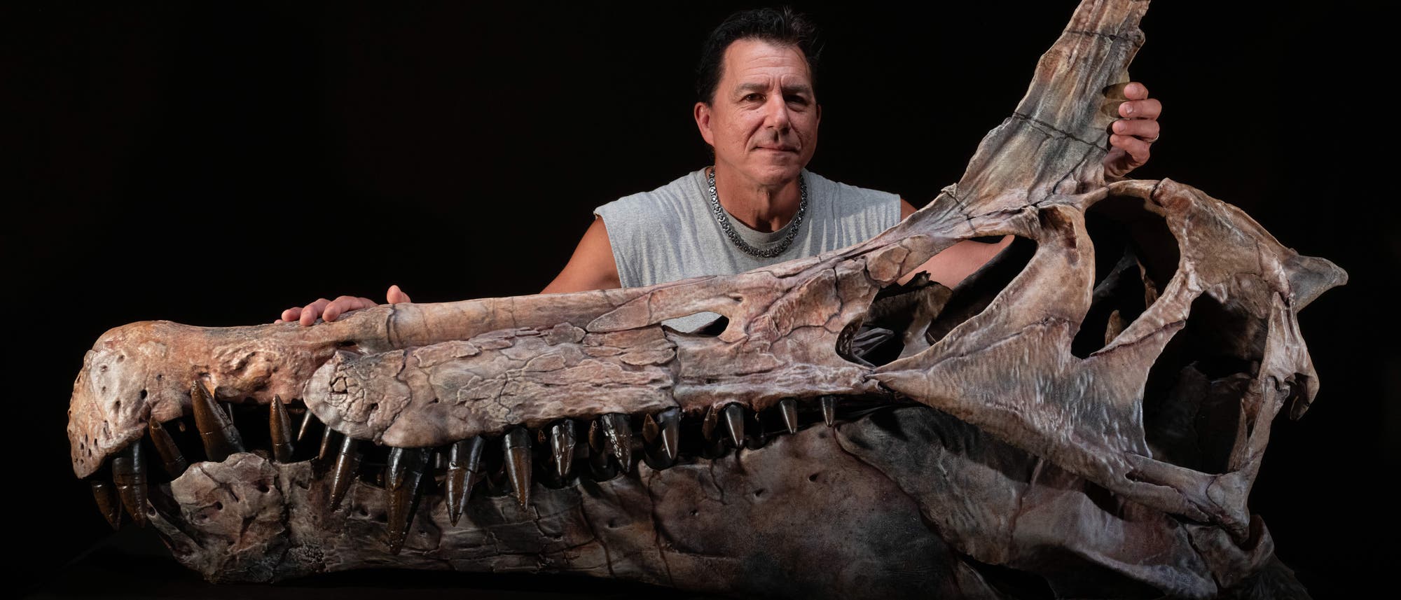 Eine Person steht hinter einem gro&szlig;en Dinosauriersch&auml;del, der auf einem Tisch liegt. Der Sch&auml;del hat lange, scharfe Z&auml;hne und eine markante, gebogene Schnauze. Die Person h&auml;lt den Sch&auml;del mit beiden H&auml;nden und blickt in die Kamera. Der Hintergrund ist dunkel, wodurch der Sch&auml;del und die Person hervorgehoben werden.